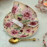 Left side view of pink butter rose floral tasseography teacup tilted on matching saucer with gold crystal teaspoon beside it