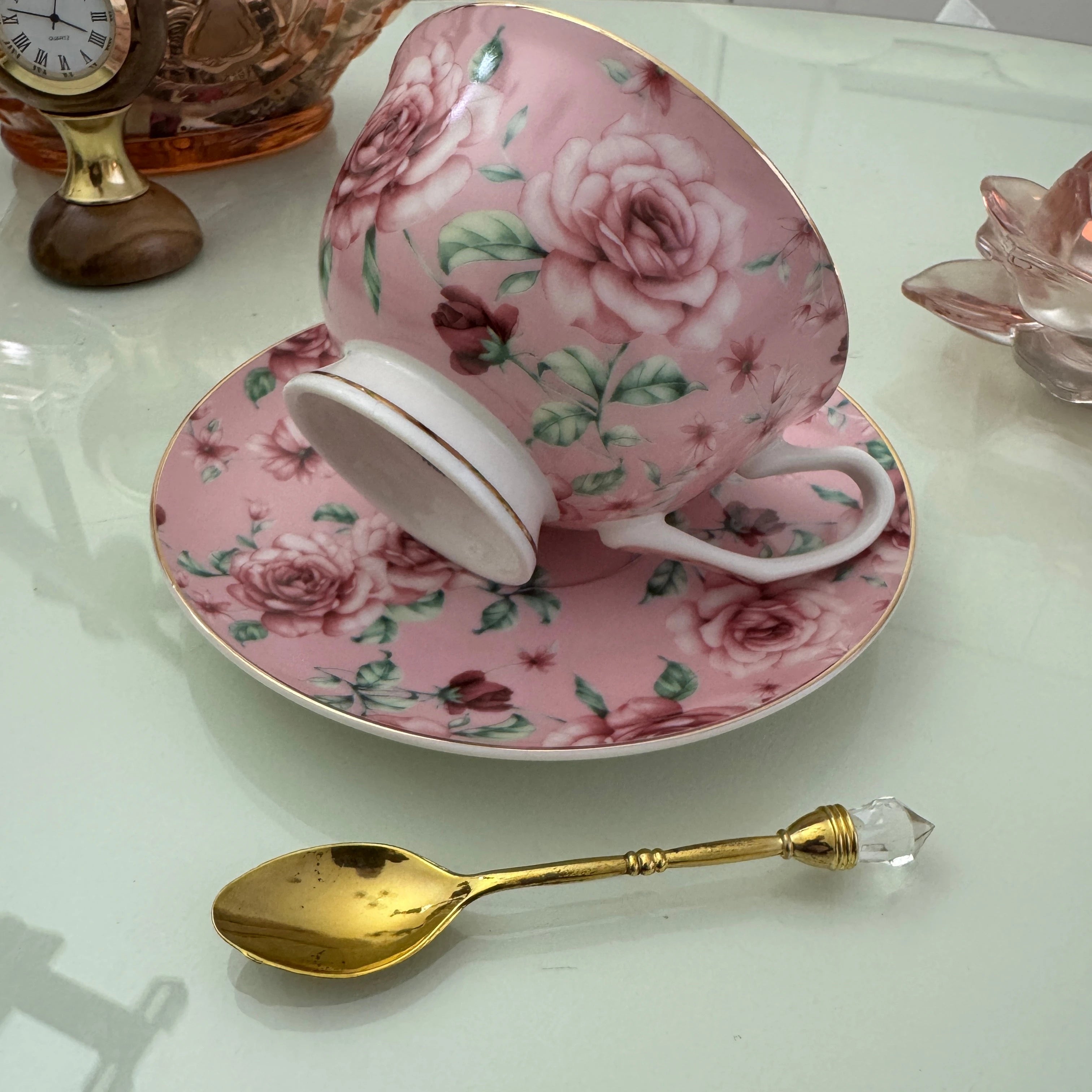 Left side view of pink rose floral tasseography teacup tilted on matching saucer with gold crystal teaspoon beside it