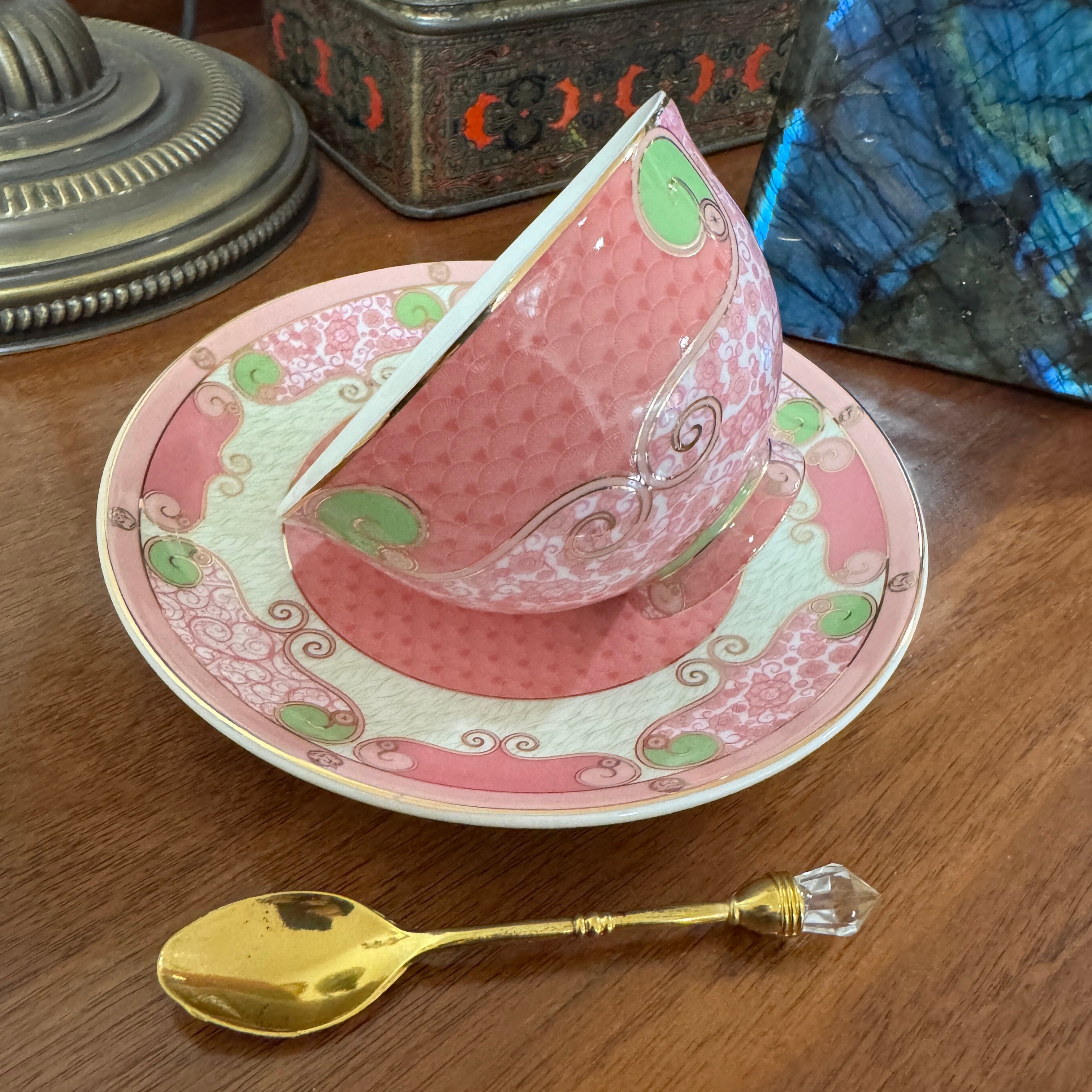 Decorative pink teacup and saucer with green accents on a wooden surface, accompanied by a gold spoon.