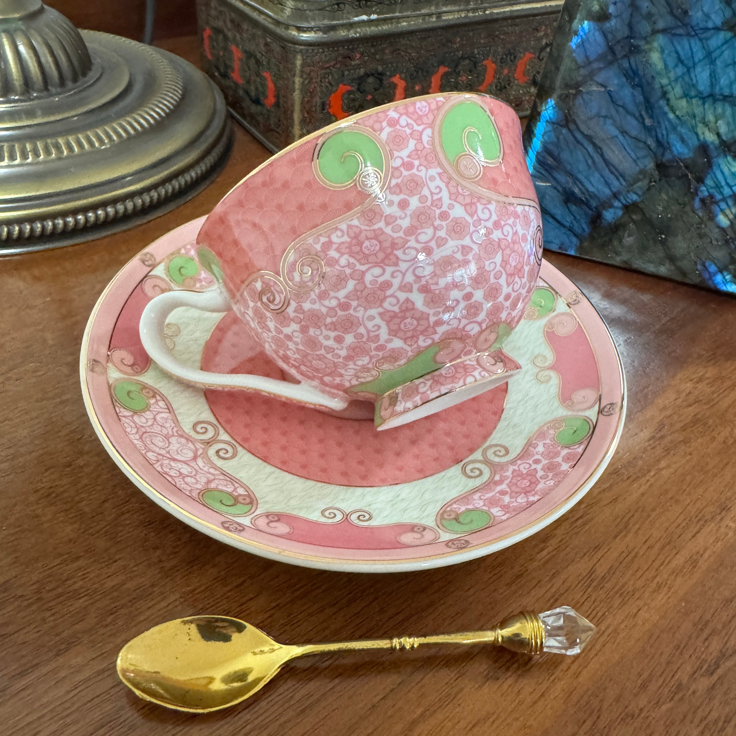 Pink teacup and saucer with green accents on a wooden surface, accompanied by a gold spoon.