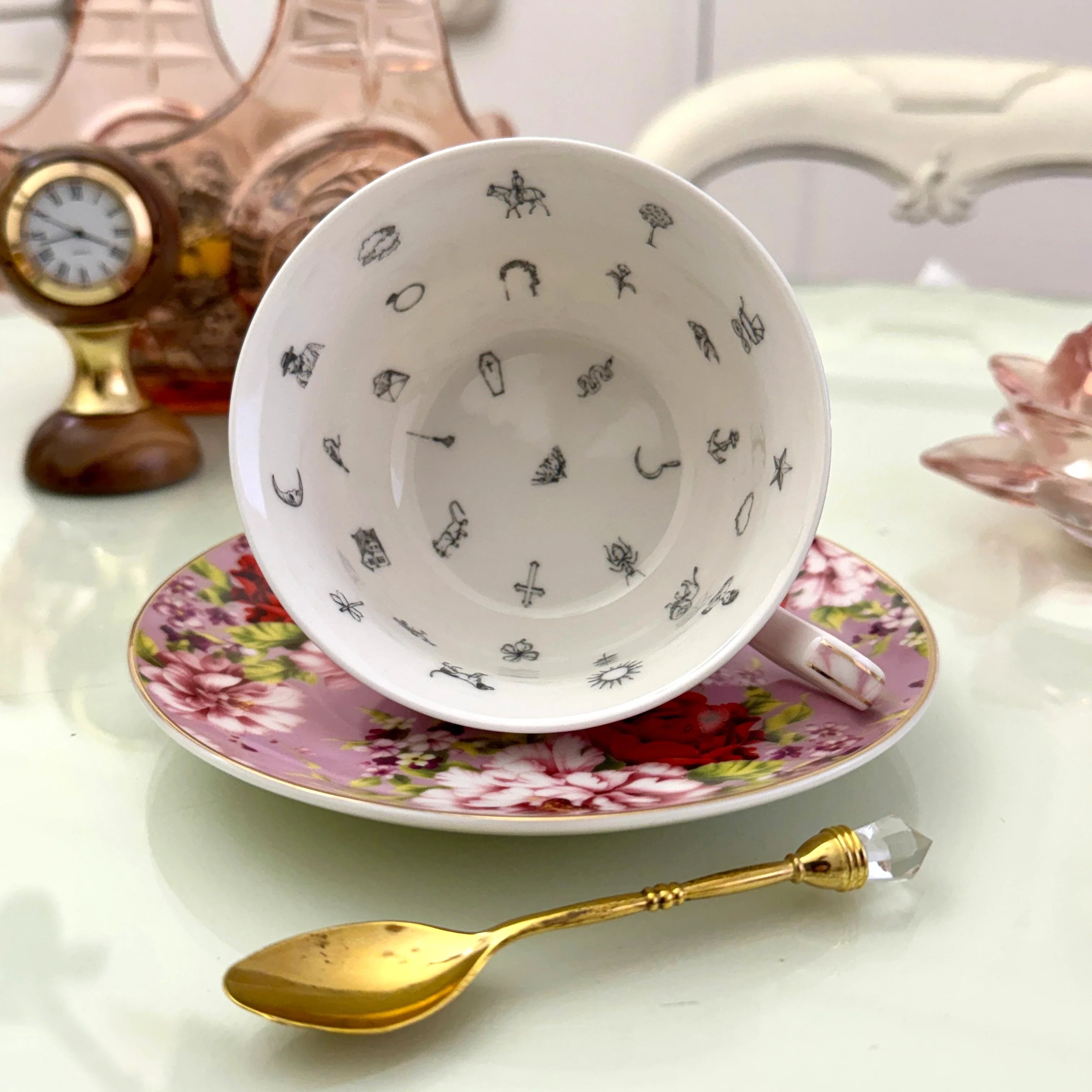 Inside view of pink red rose floral tasseography teacup face-up showing all 32 printed tea leaf reading divination symbols, resting on matching saucer with gold crystal teaspoon