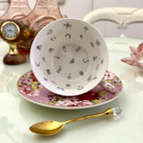 Inside view of pink red rose floral tasseography teacup face-up showing all 32 printed tea leaf reading divination symbols, resting on matching saucer with gold crystal teaspoon