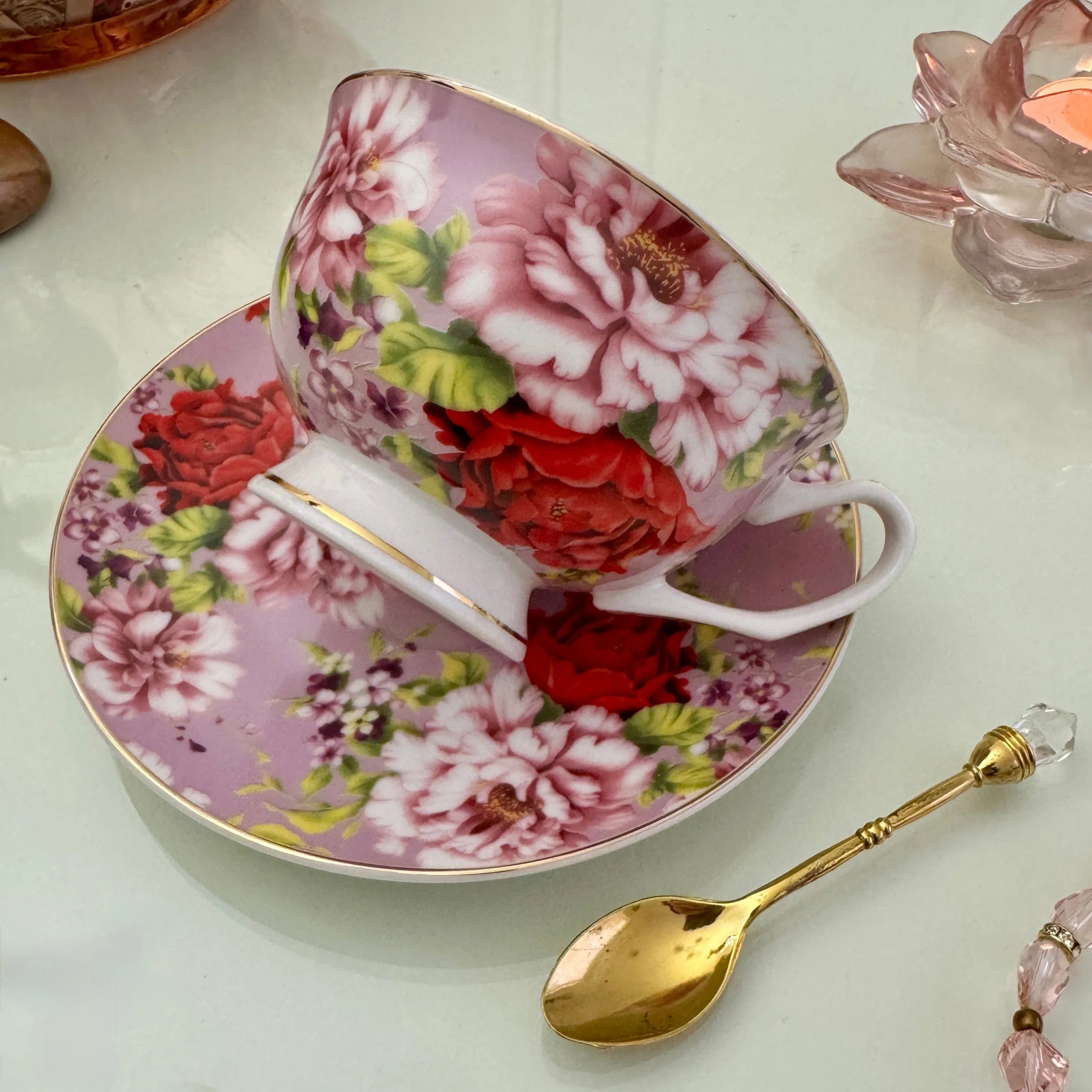 Left side view of pink red rose floral tasseography teacup tilted on matching saucer with gold crystal teaspoon beside it