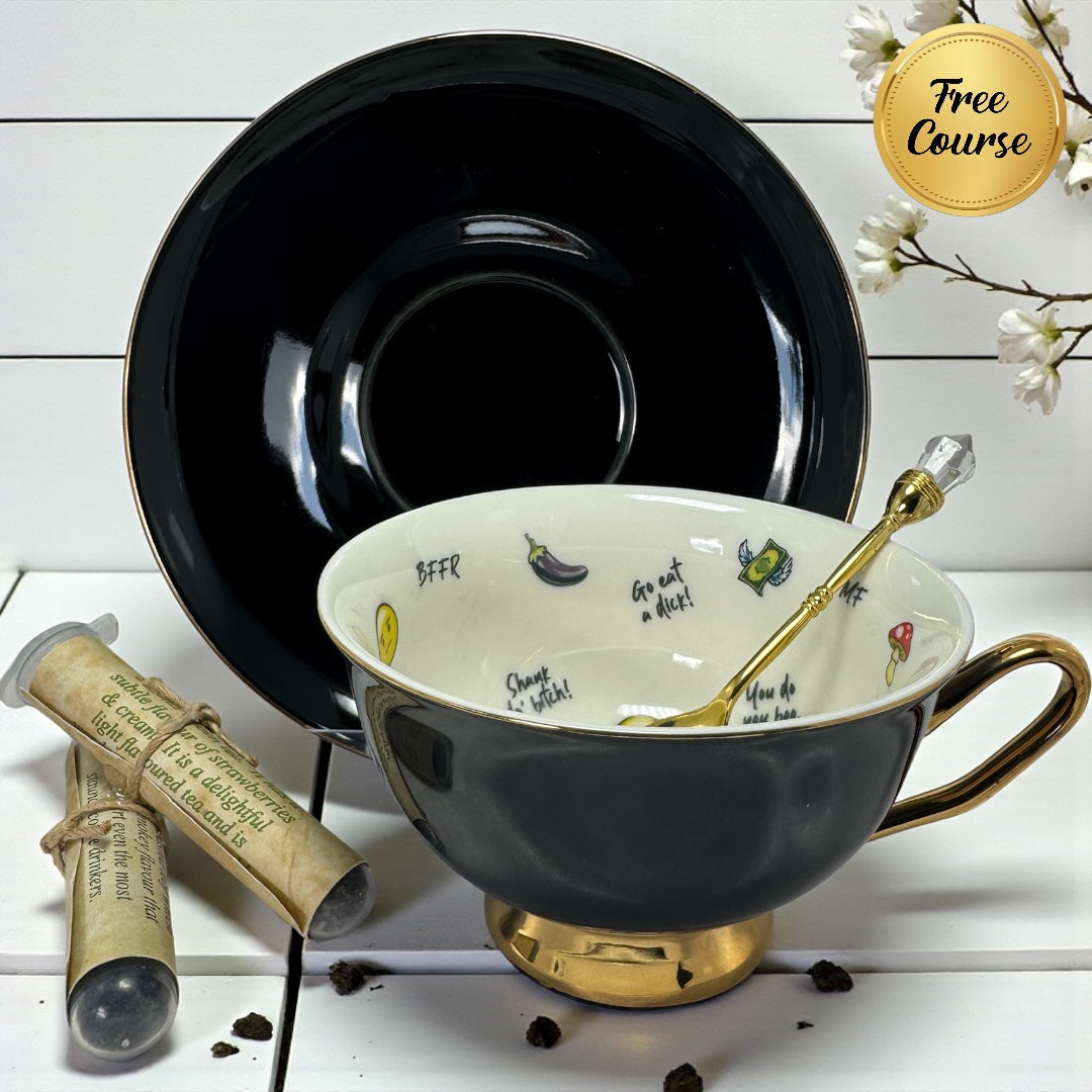 Black and gold teacup and saucer with internal irreverent emojis and text, a gold crystal spoon, and two tea sample tubes.