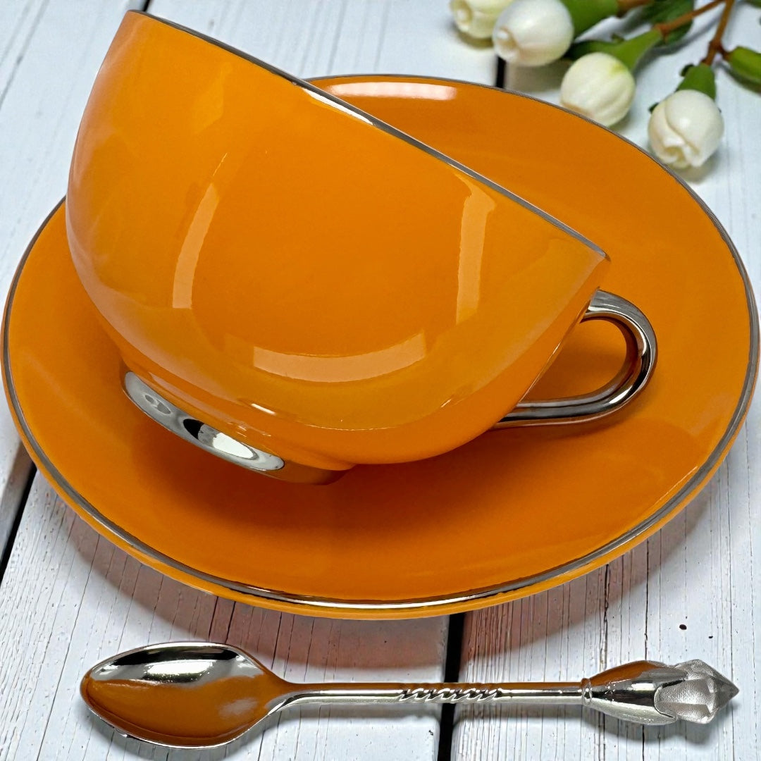 Side profile of a glossy orange tea cup with a silver handle sitting on a matching saucer with white flowers in the background.