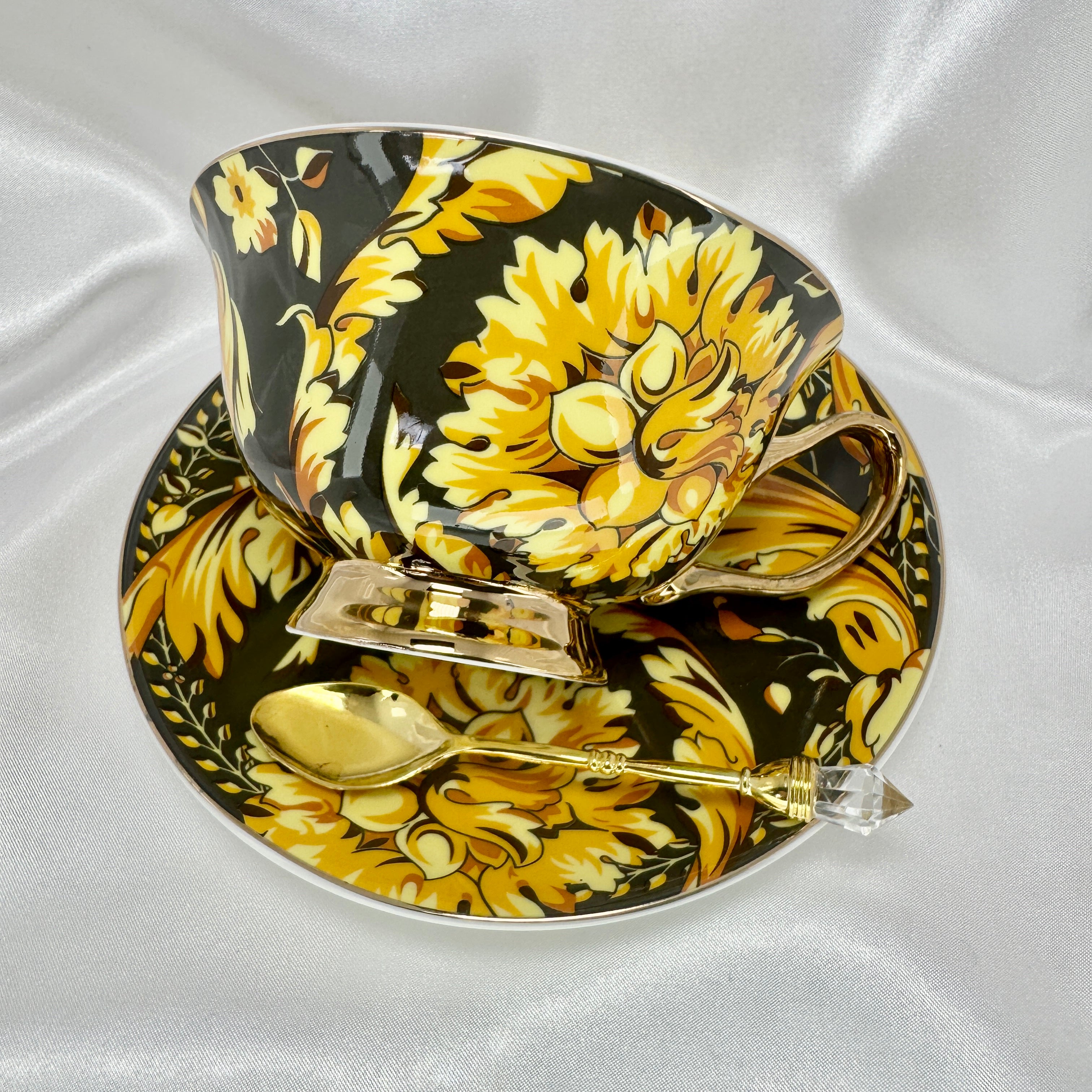 Right side view of the black and gold teacup and saucer set with a gold handle and spoon, resting on white satin