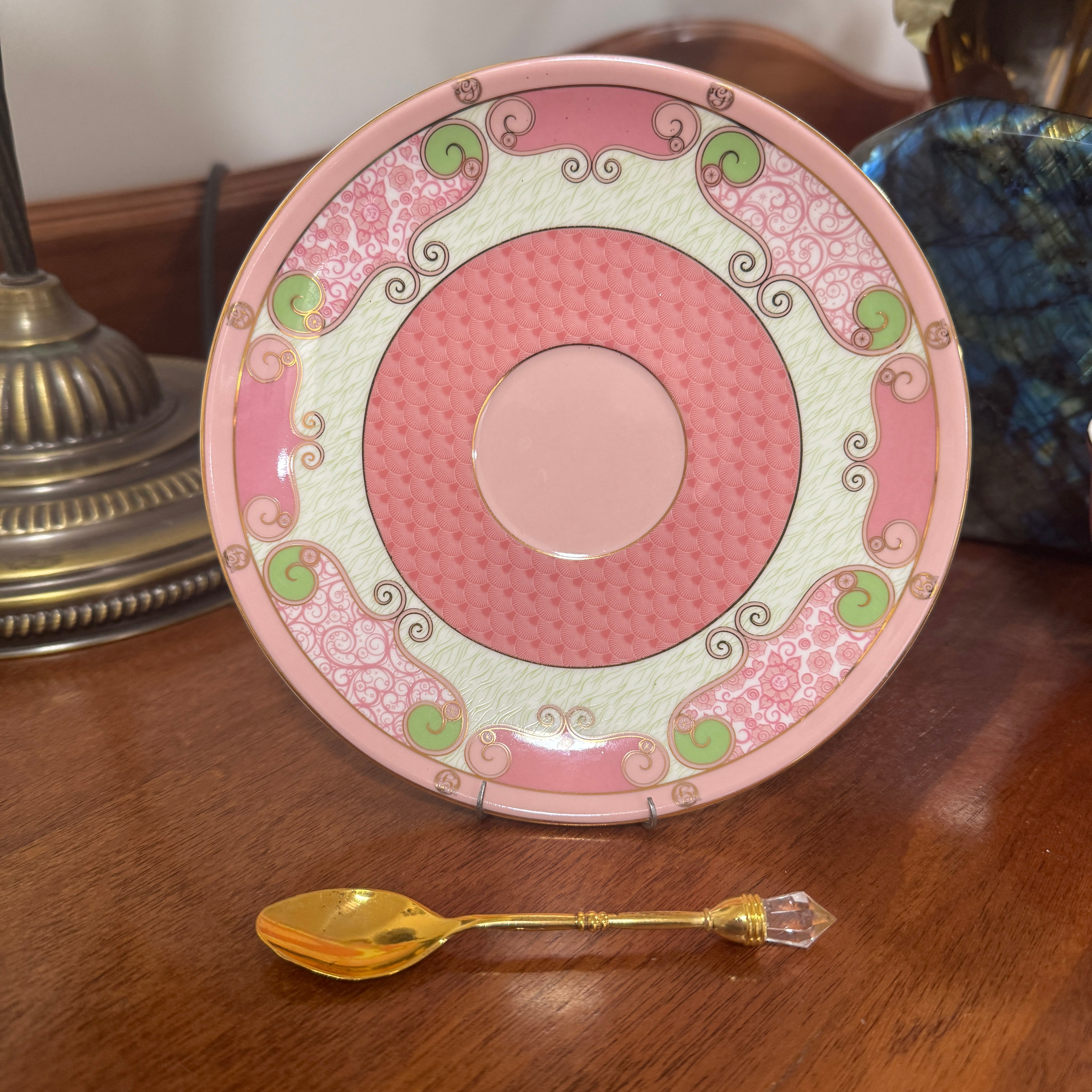 Decorative pink plate with green patterns on a wooden surface next to a gold spoon.
