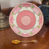 Decorative pink plate with green patterns on a wooden surface next to a gold spoon.