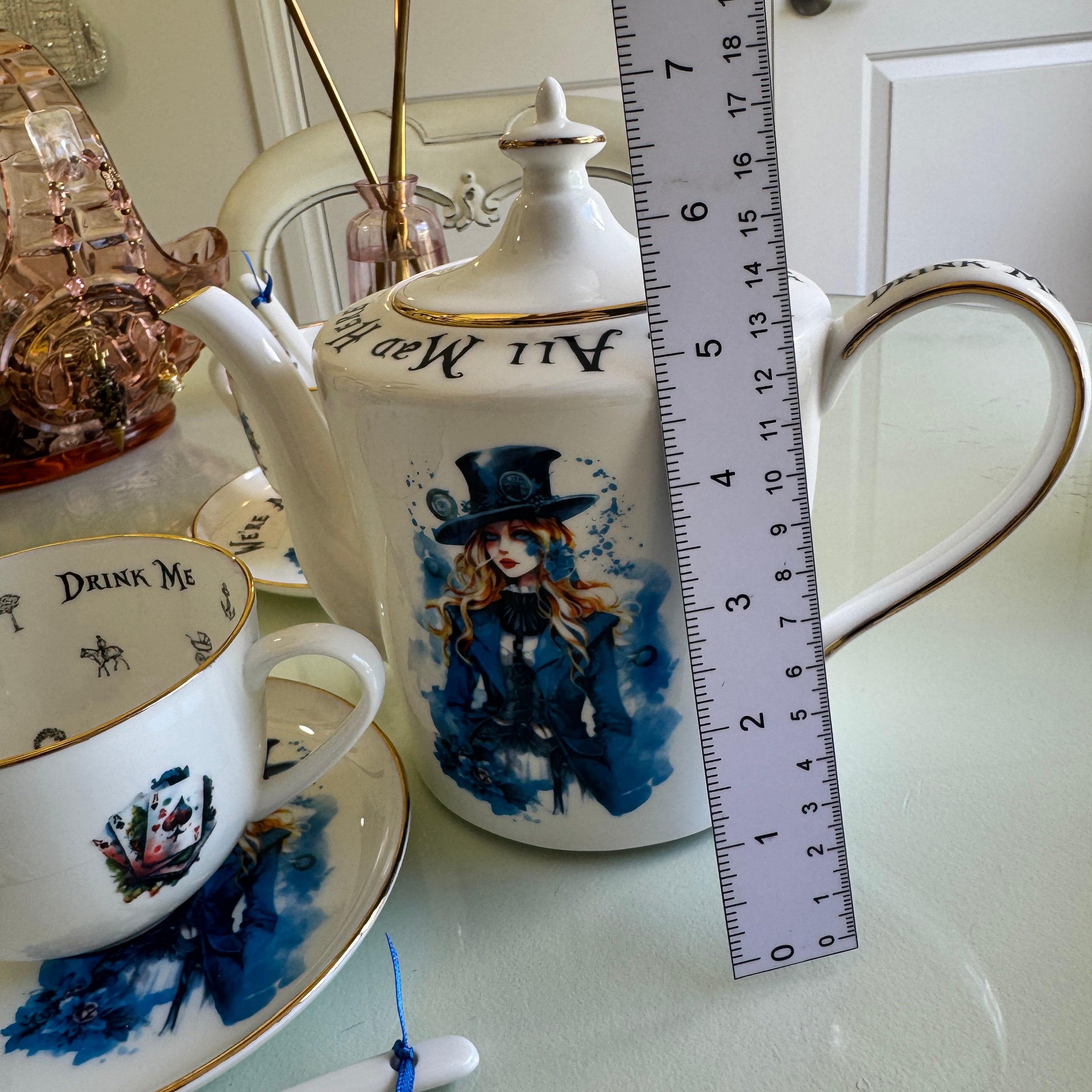 Teapot and teacup with 'Drink Me' and 'All We Need' text, next to a ruler for scale.