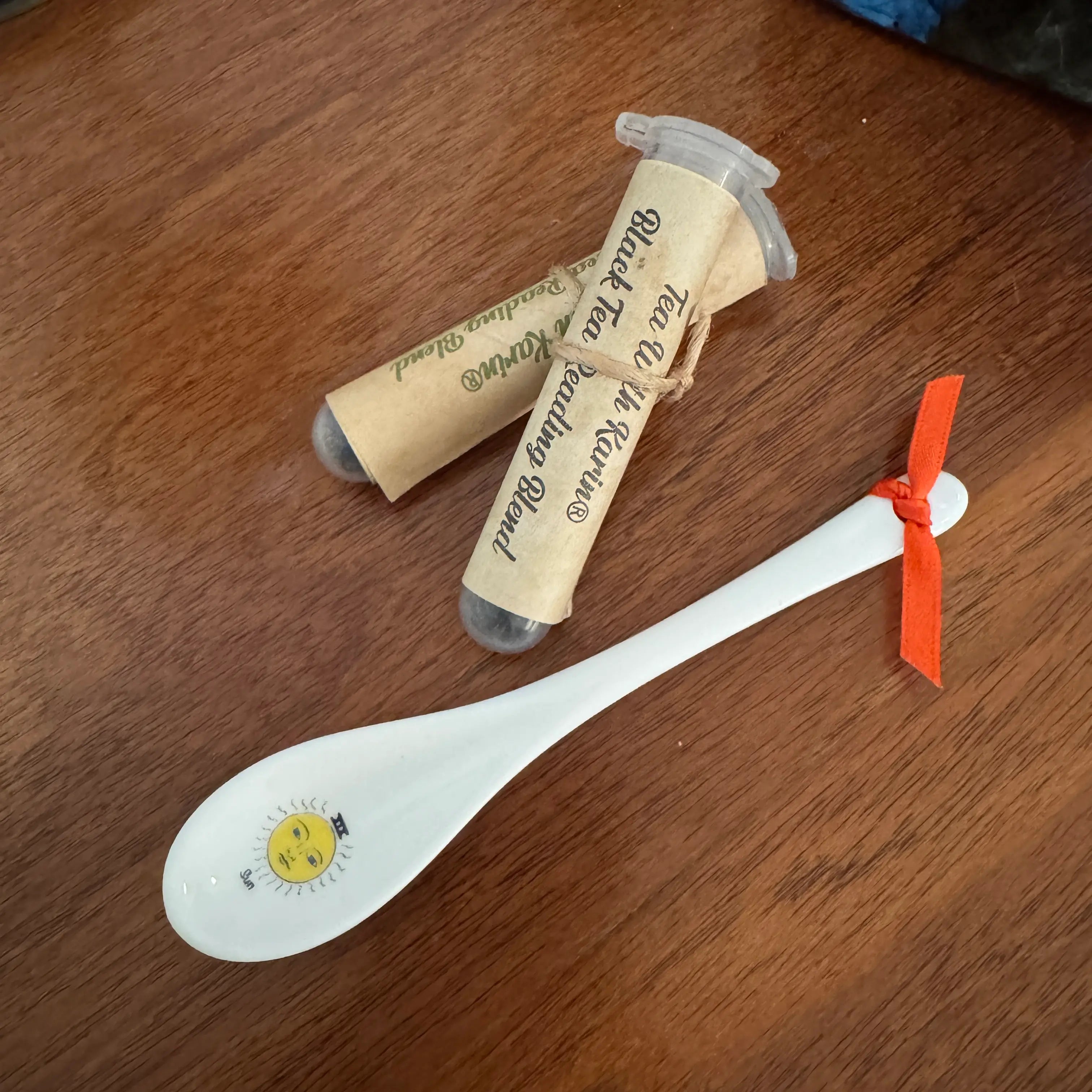 A white ceramic tea spoon with a yellow sun illustration on the bowl, tied with an orange ribbon, displayed next to two rolled kraft paper tubes labeled 'Tea With Me' on a wooden tabletop.