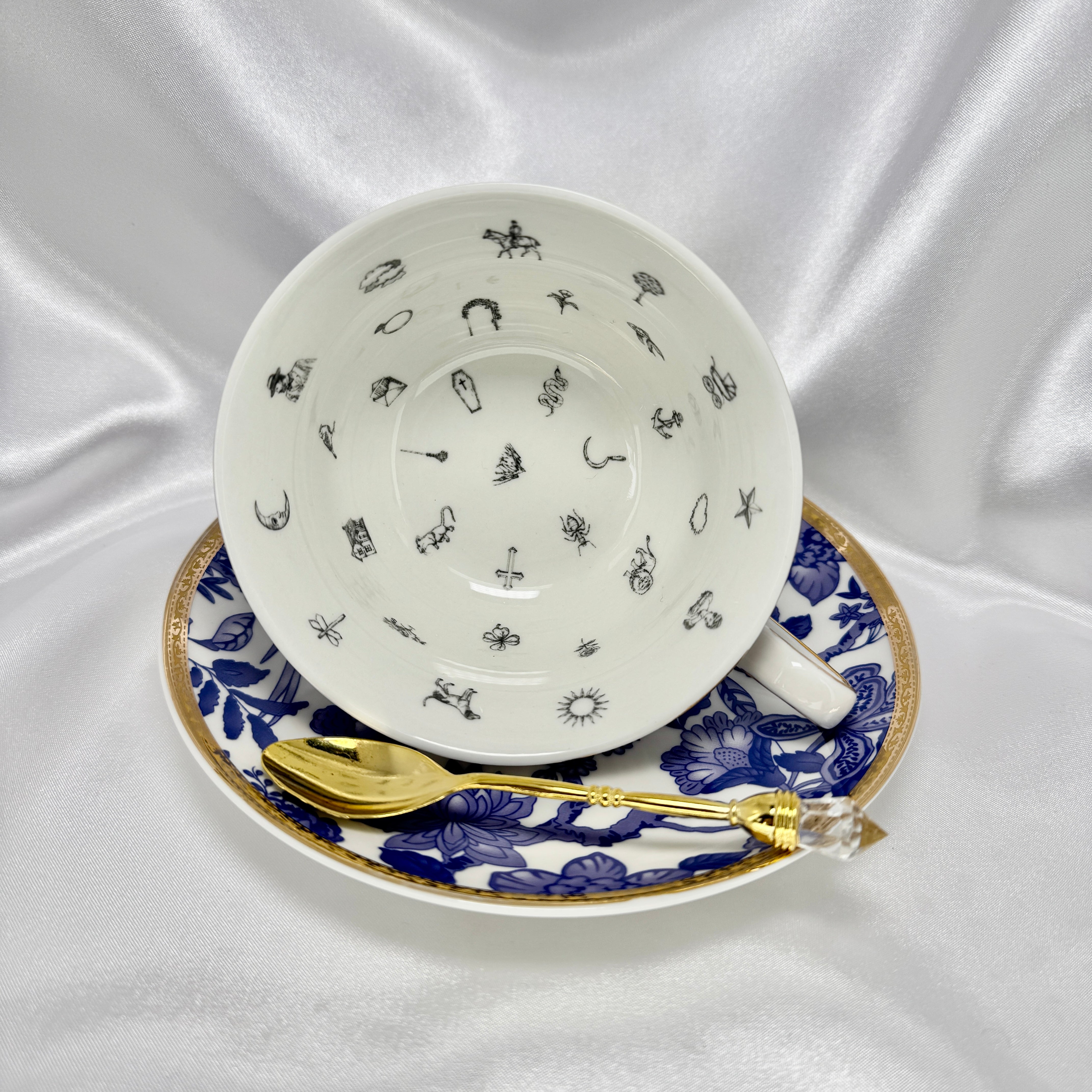 Blue and white teacup and saucer set with gold trim, cup tilted on its side showing tiny black divination symbols inside.