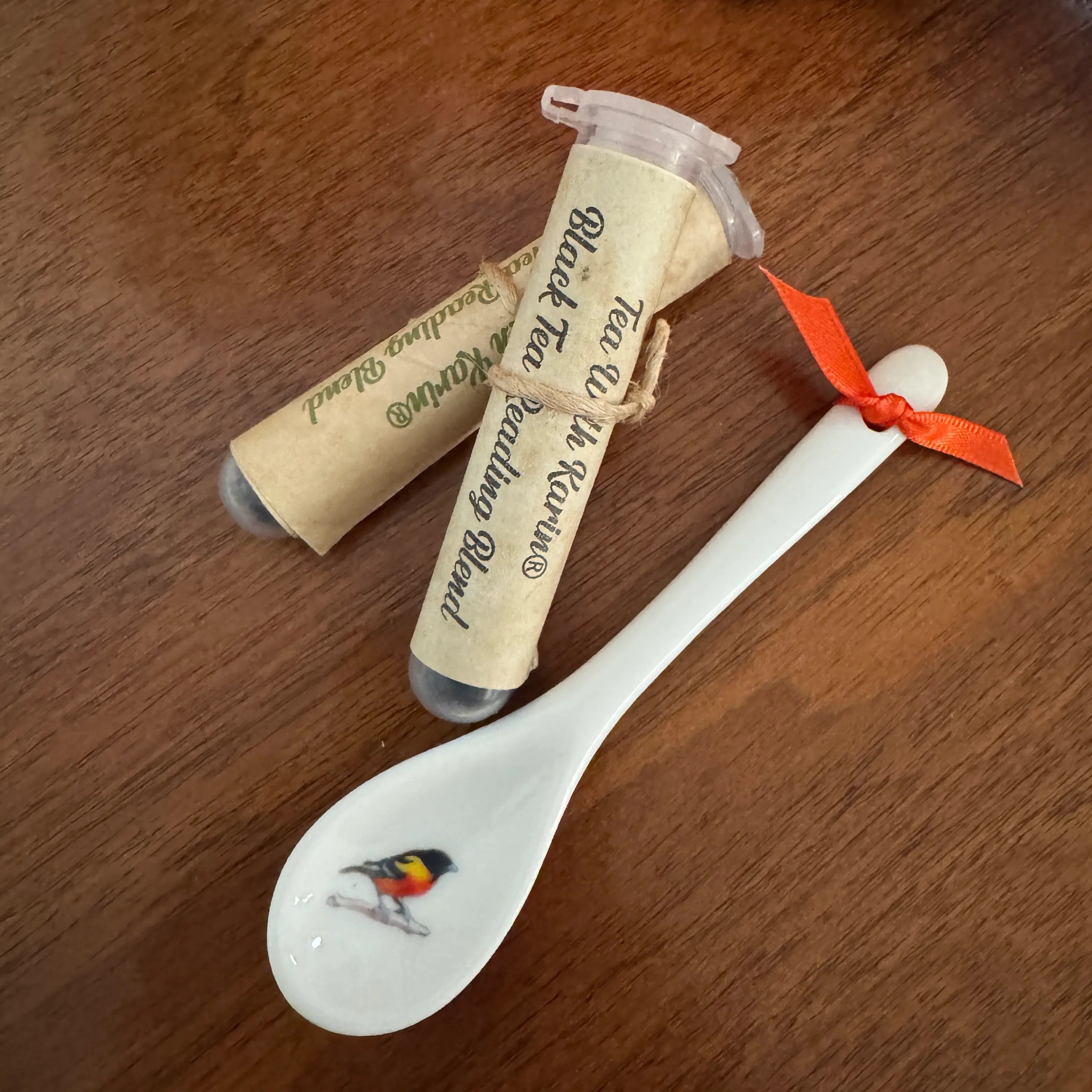A white ceramic tea spoon with a small bird illustration in the bowl, tied with an orange ribbon, next to two rolled parchment-style tea bags labeled 'Black Tea Mixture Blend' on a wooden surface.