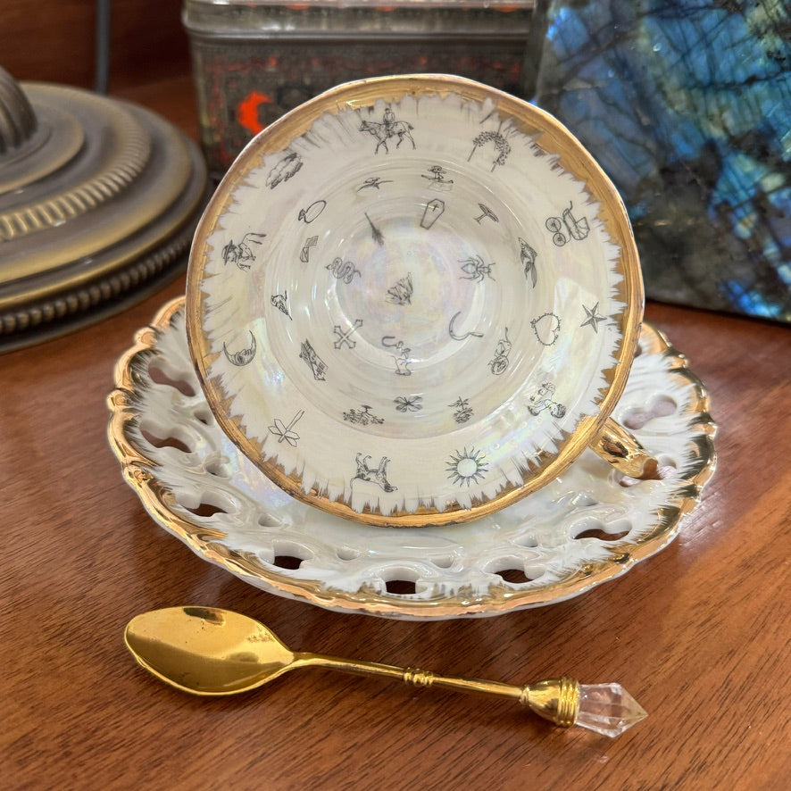 Decorative teacup with gold rim and saucer on a wooden surface, accompanied by a gold spoon.