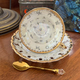 Decorative teacup with gold rim and saucer on a wooden surface, accompanied by a gold spoon.