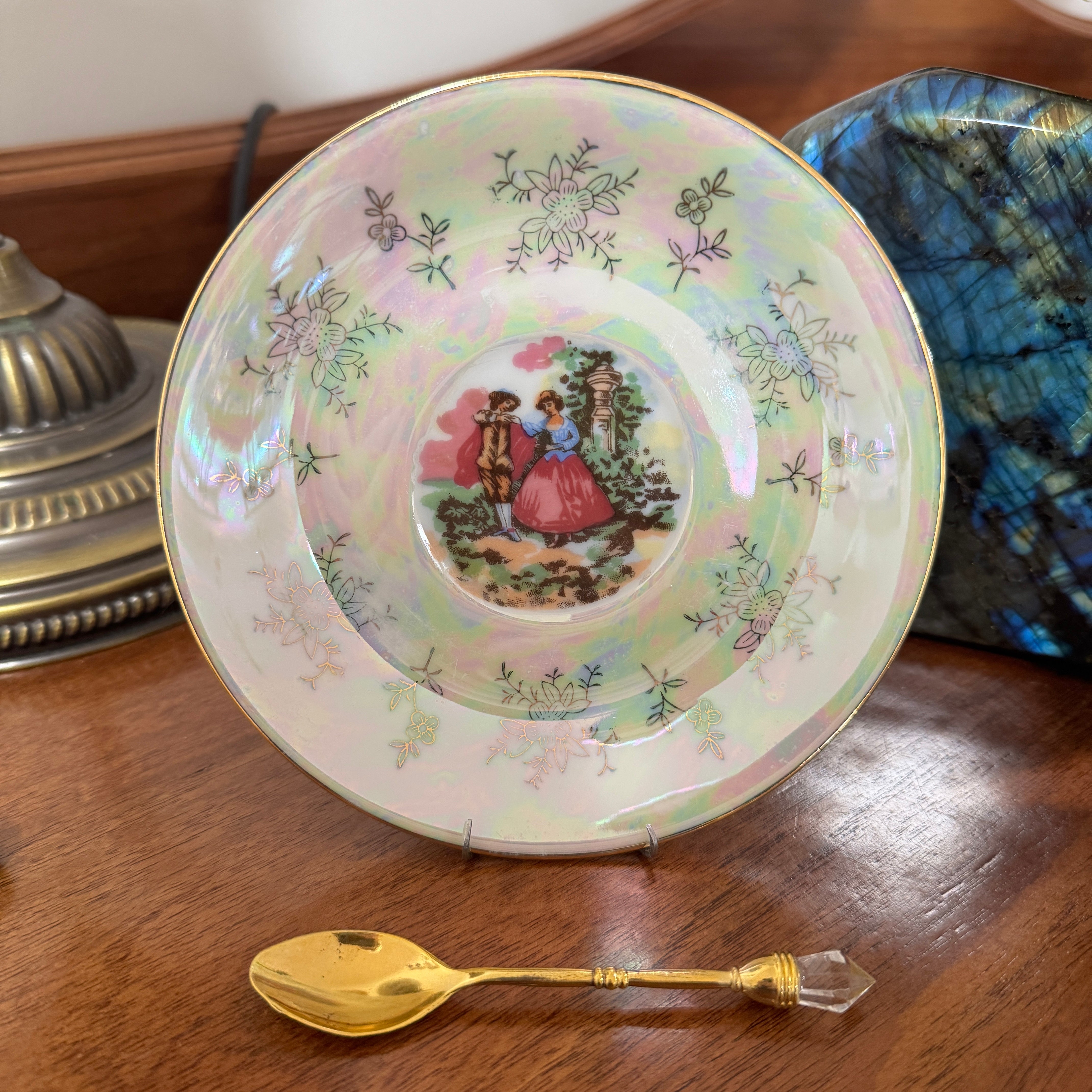Decorative plate with a vintage illustration of a couple in a garden, placed on a wooden surface with a gold spoon.