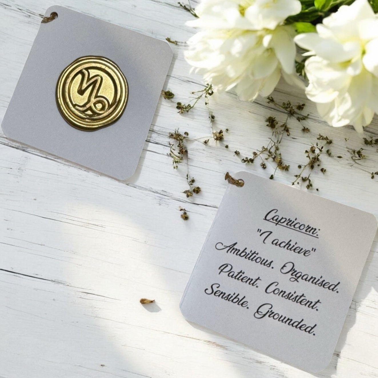 Two cards on a wooden surface with flowers, one featuring a gold emblem and the other with text about Capricorn.