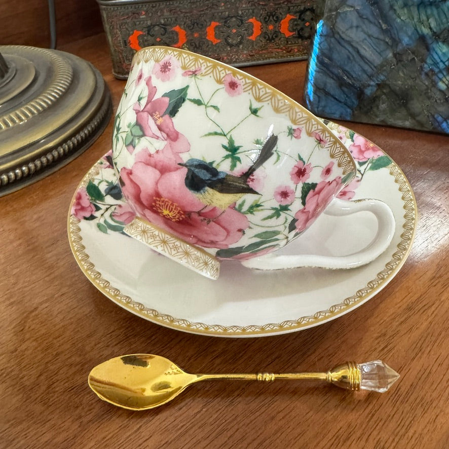 Decorative teacup with floral design and bird, placed on a matching saucer with a gold spoon beside it on a wooden surface.