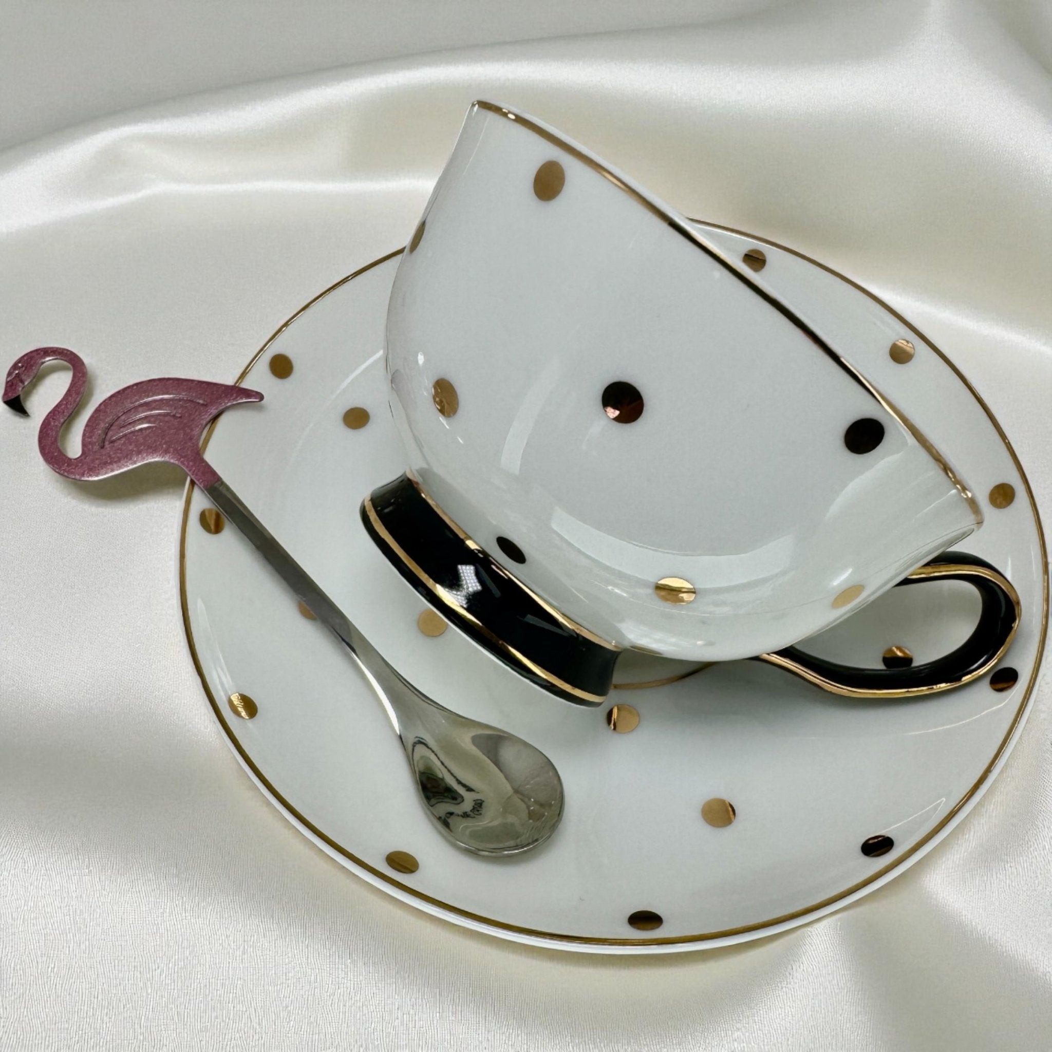 Elegant white new bone china tea cup and saucer set with gold and black polka dots. Close-up showing the metallic pink flamingo stirring spoon resting against the saucer.