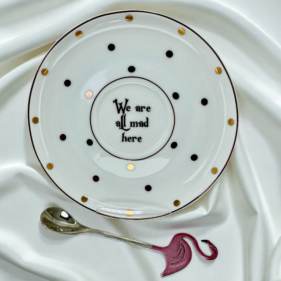 New bone china tea saucer with gold and black polka dots, featuring the quote 'We are all mad here' in the center. Shown next to a polished silver spoon and a decorative pink flamingo handle.