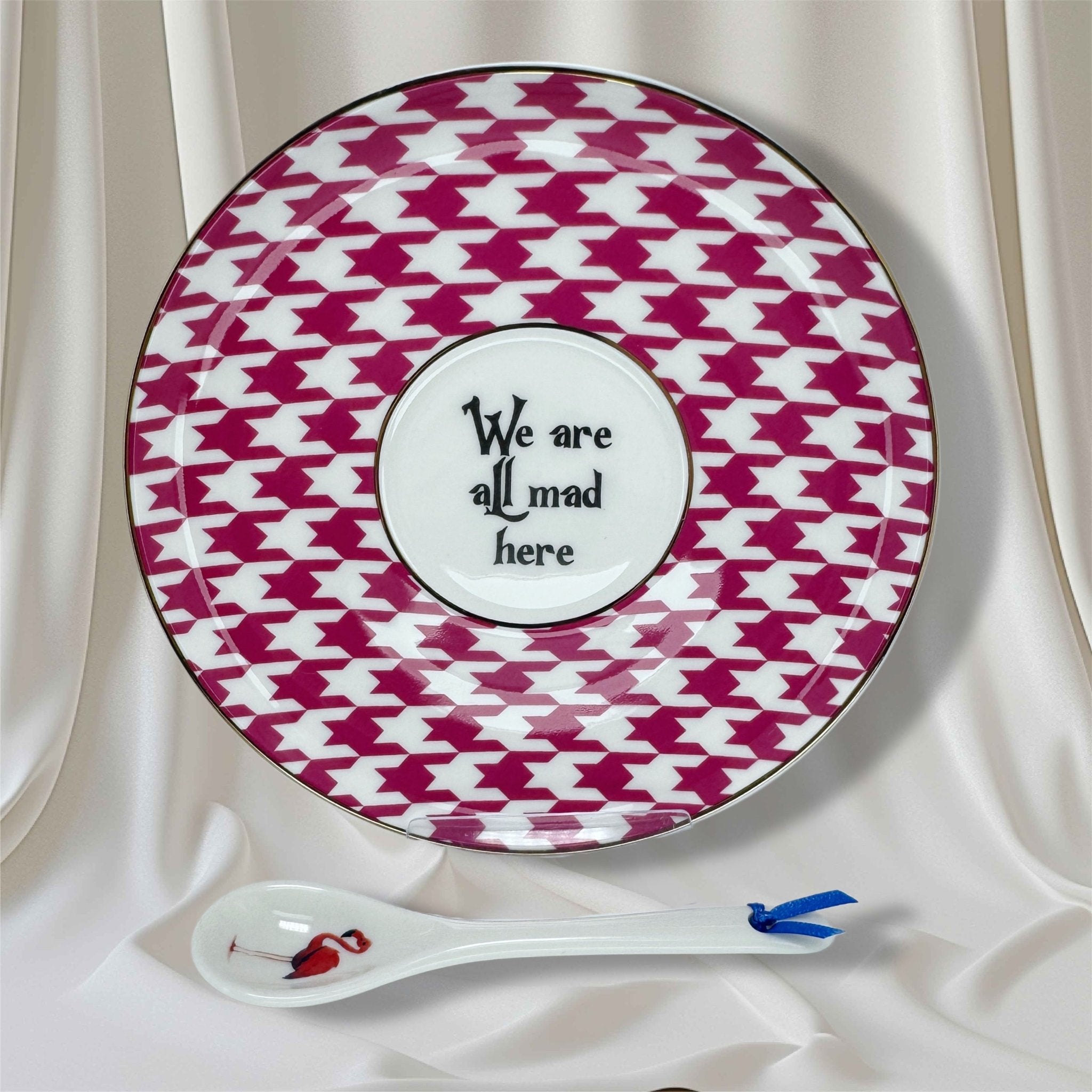Decorative plate with pink and white pattern and 'We are all mad here' text, placed on a white surface.