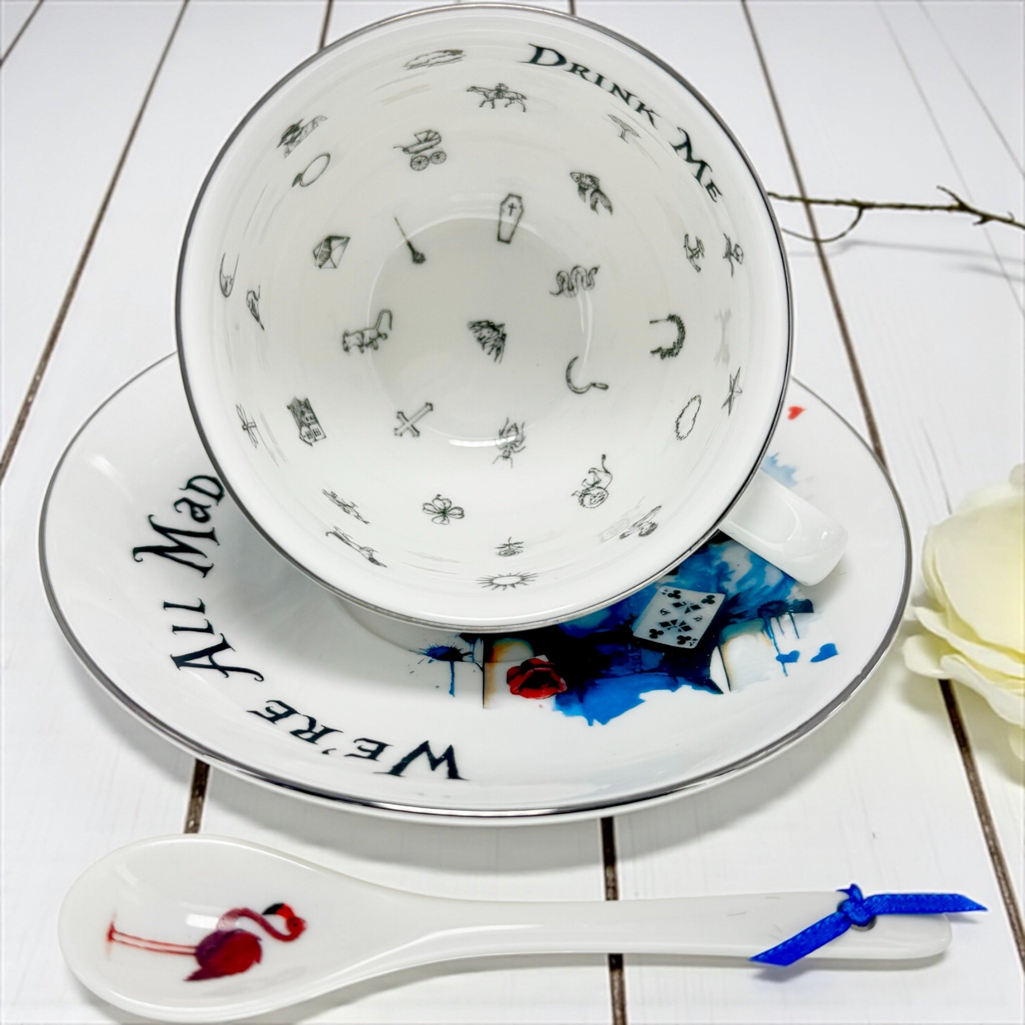 Teacup with 'Drink Me' and 'We're All Mad Here' text, saucer, and spoon on a white wooden surface.