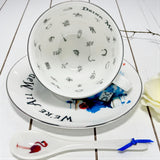 Teacup with 'Drink Me' and 'We're All Mad Here' text, saucer, and spoon on a white wooden surface.