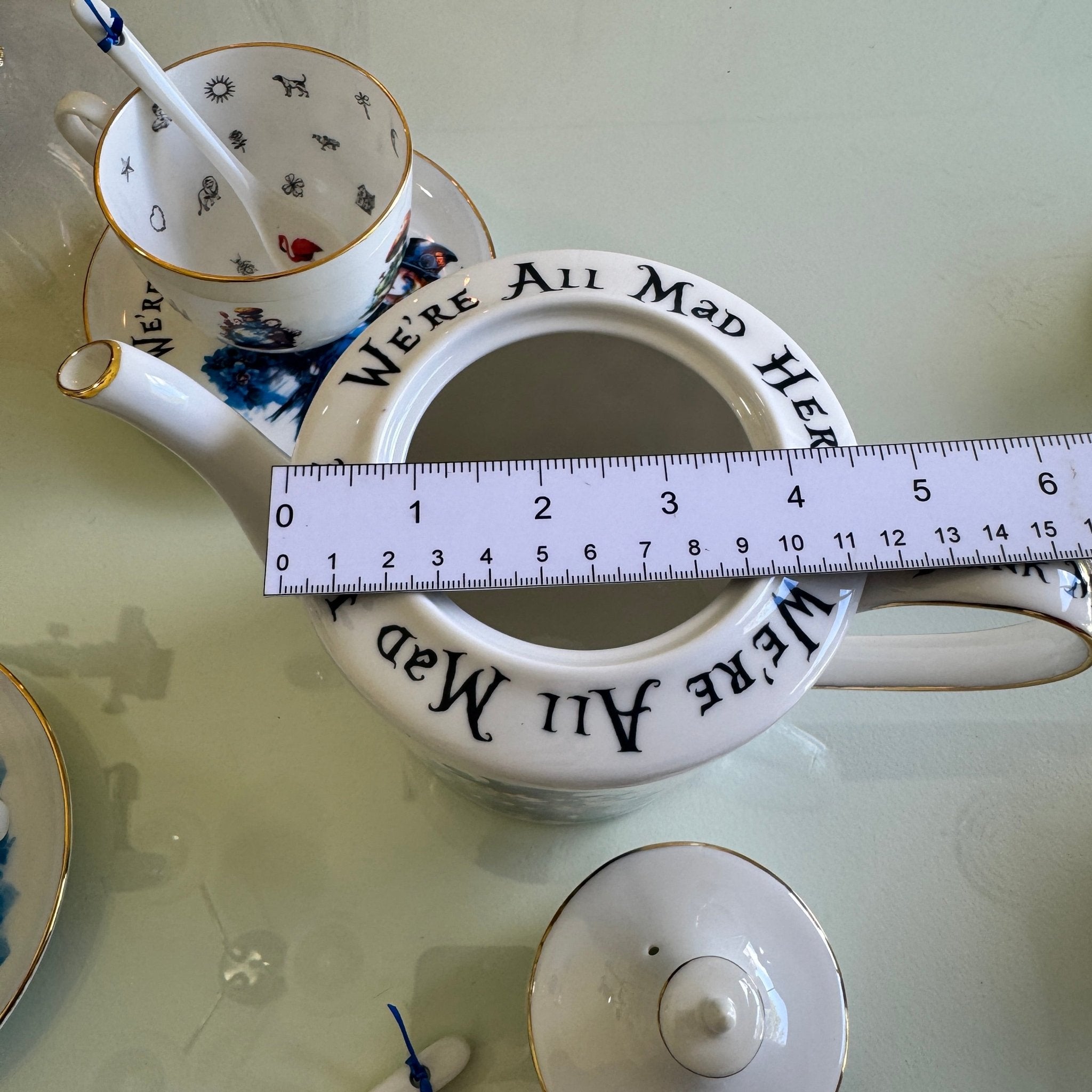 Teapot with 'We're All Mad Here' text, a measuring tape, and a spoon on a light green surface.