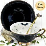 Black teacup with gold handle and spoon, white saucer with bat design, and tea leaves on a wooden surface.
