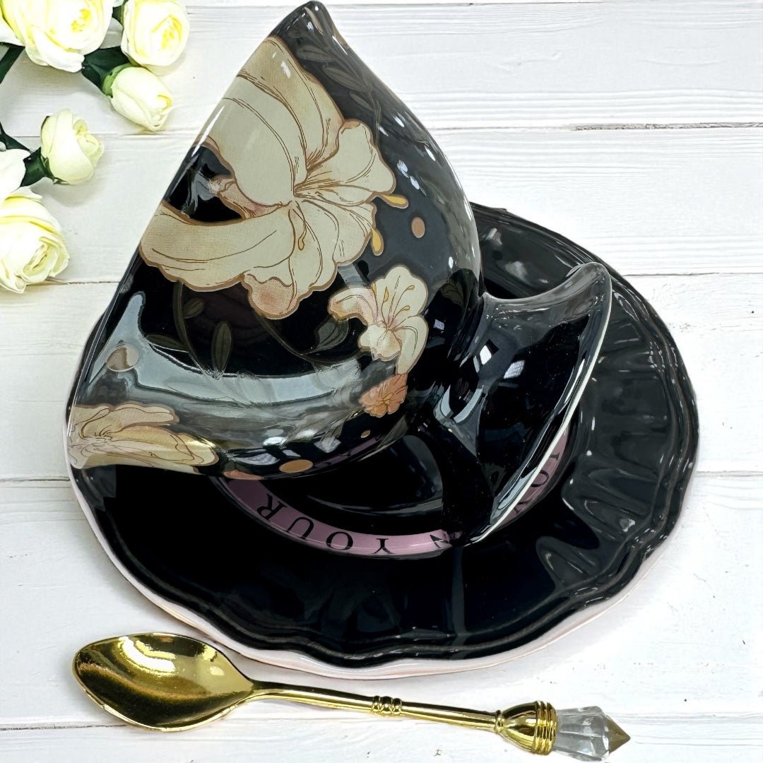 Black teacup and saucer set with floral design for tea leaf reading on a white wooden surface with gold spoon.