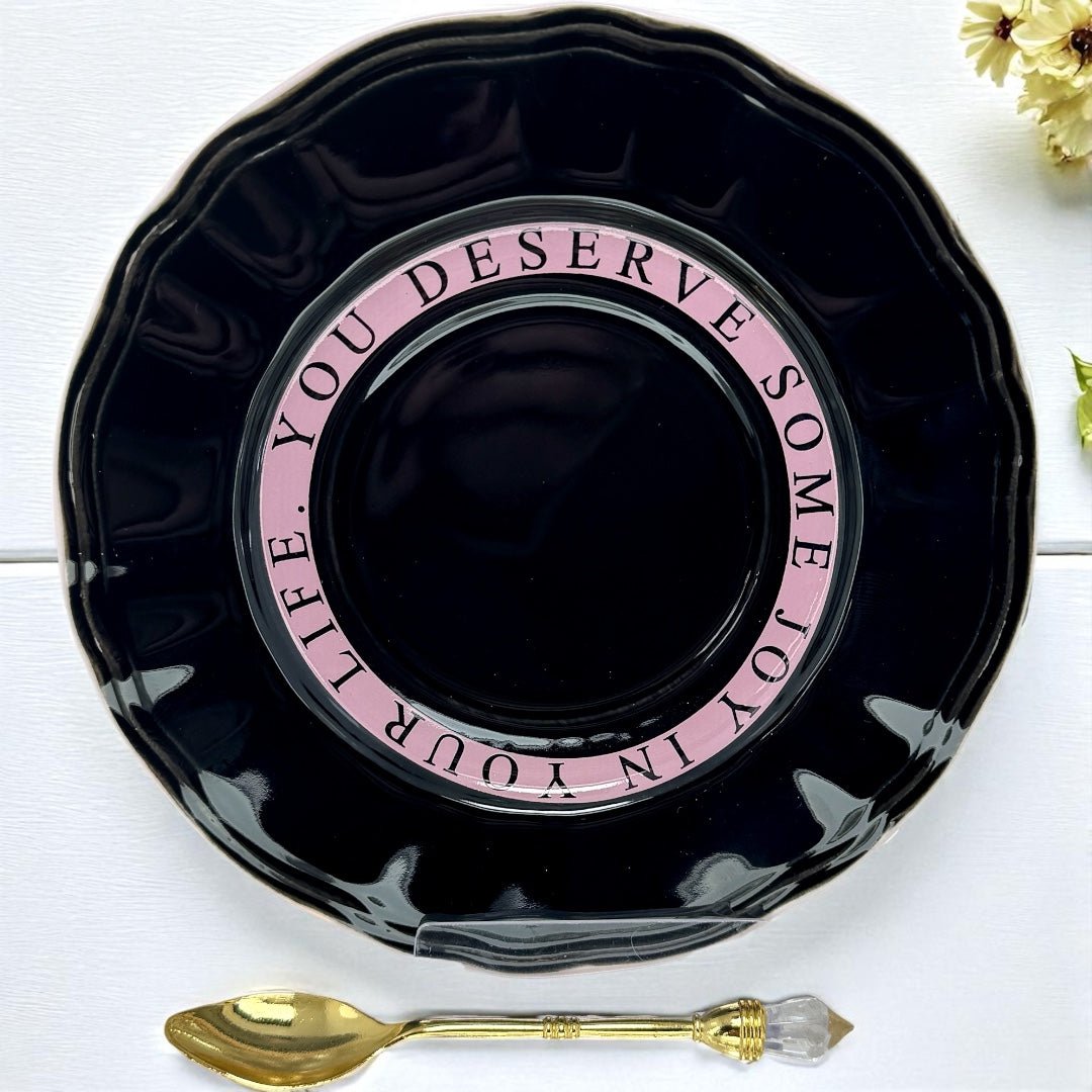 Black saucer with pink text and a gold spoon on a white surface.