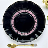 Black saucer with pink text and a gold spoon on a white surface.
