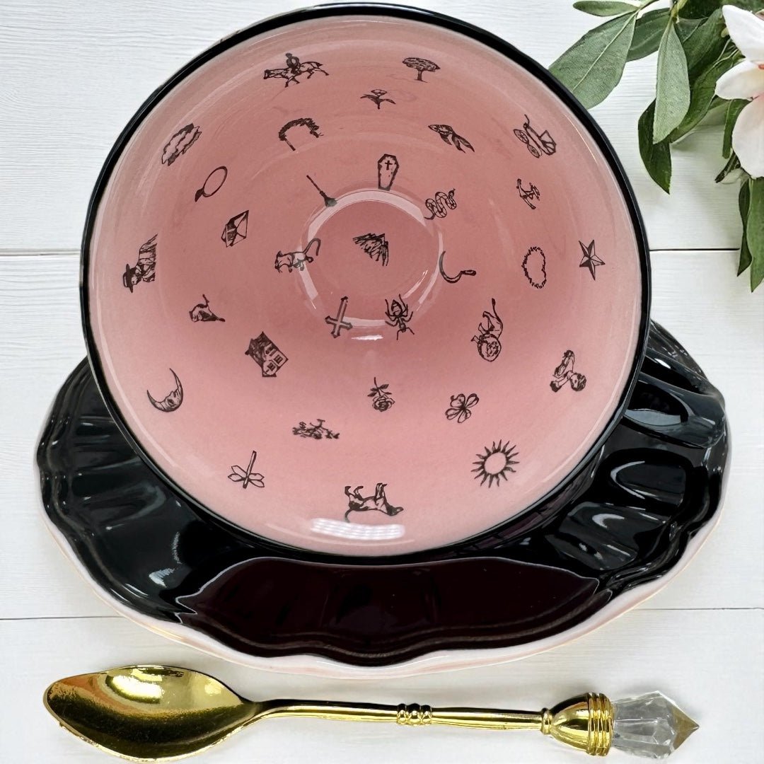Pink ceramic teacup with black designs on a white wooden surface with a gold spoon and green leaves.