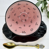 Pink ceramic teacup with black designs on a white wooden surface with a gold spoon and green leaves.
