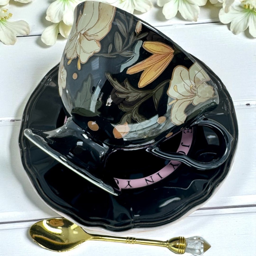 Black floral teacup and saucer set with a gold spoon on a white wooden surface.