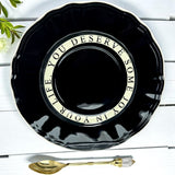 Black decorative saucer with a gold message on a white wooden surface with a gold spoon.