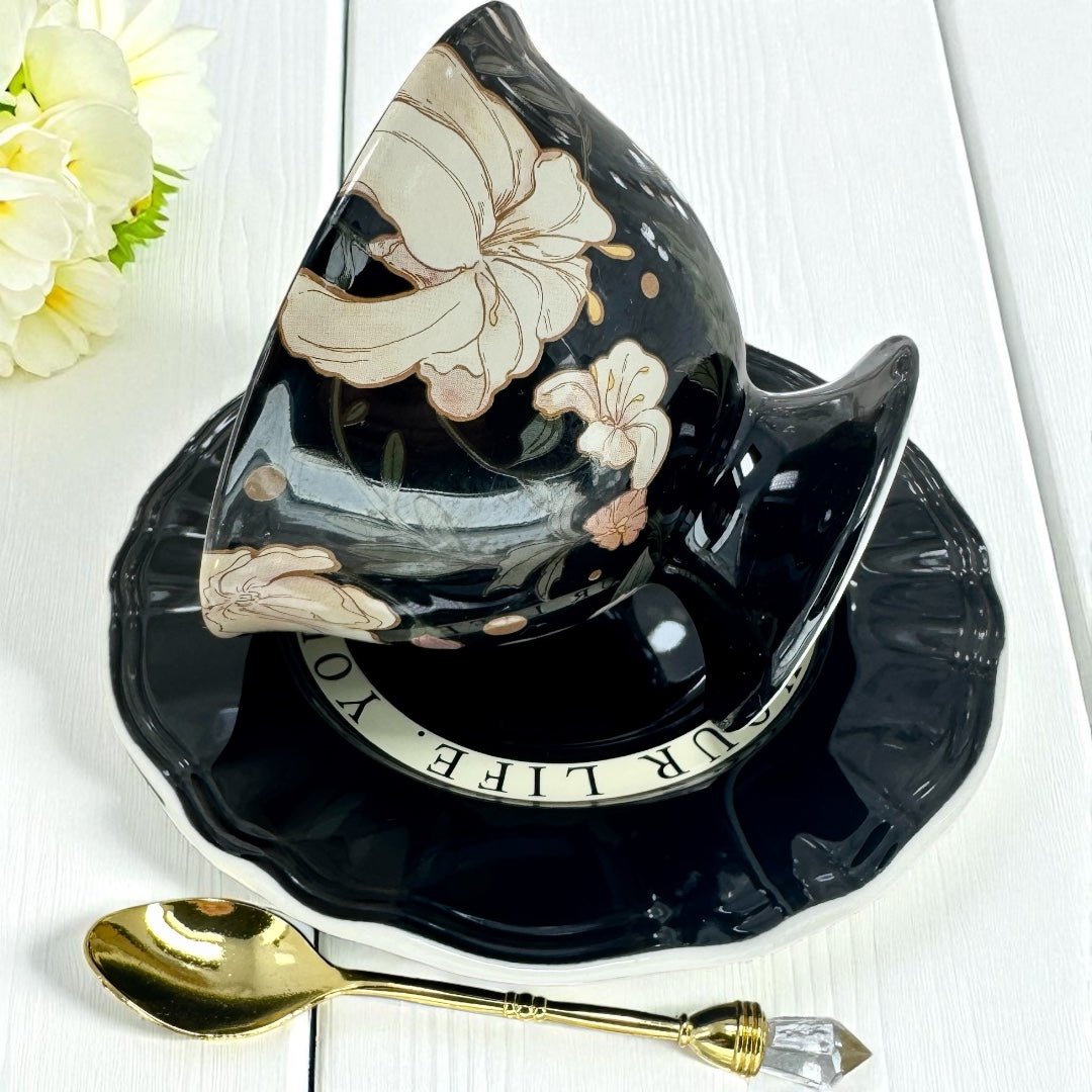Black ceramic teacup and saucer with floral design on a white surface with a gold spoon.