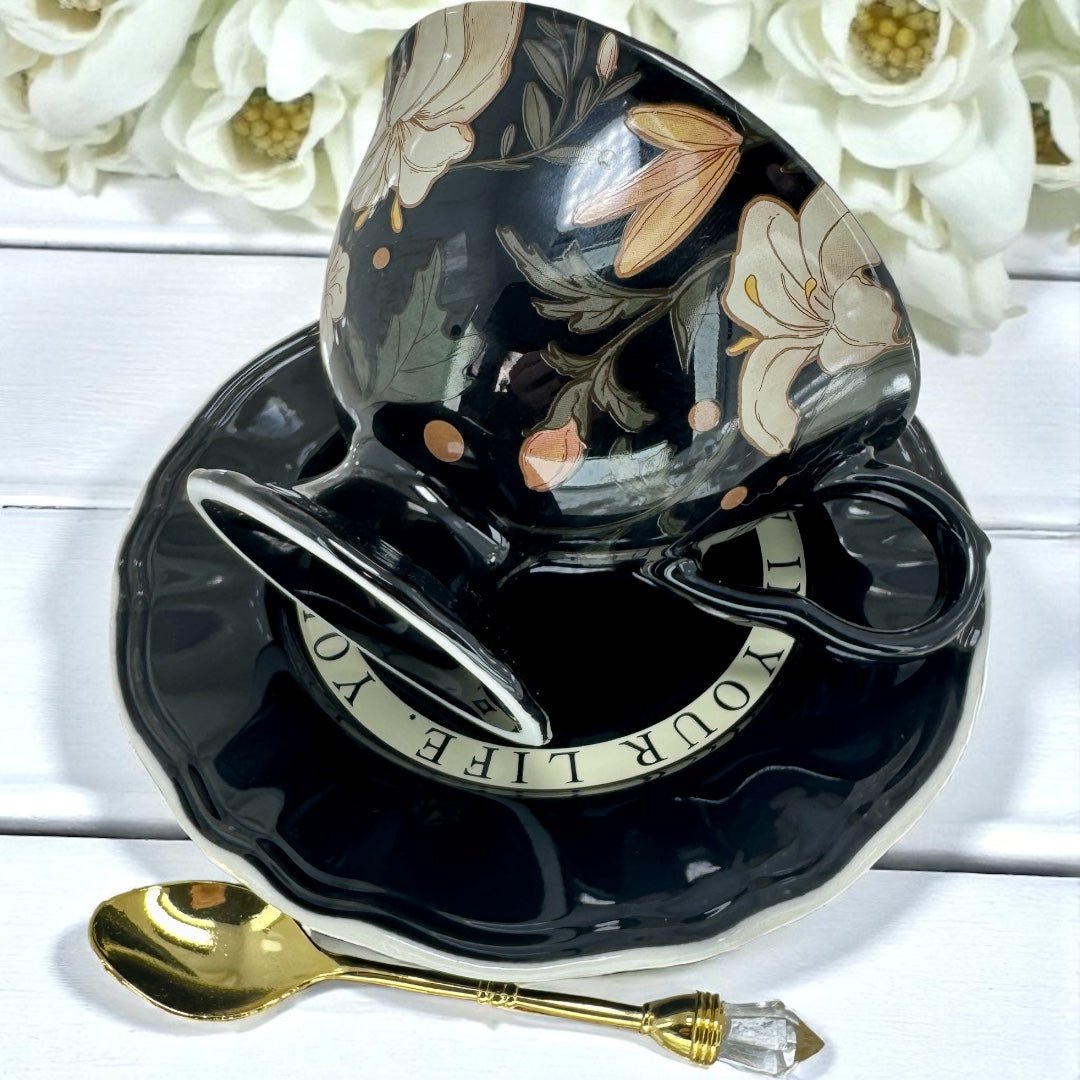 Black ceramic teacup with floral design on a saucer, accompanied by a gold spoon, on a white wooden surface with flowers.