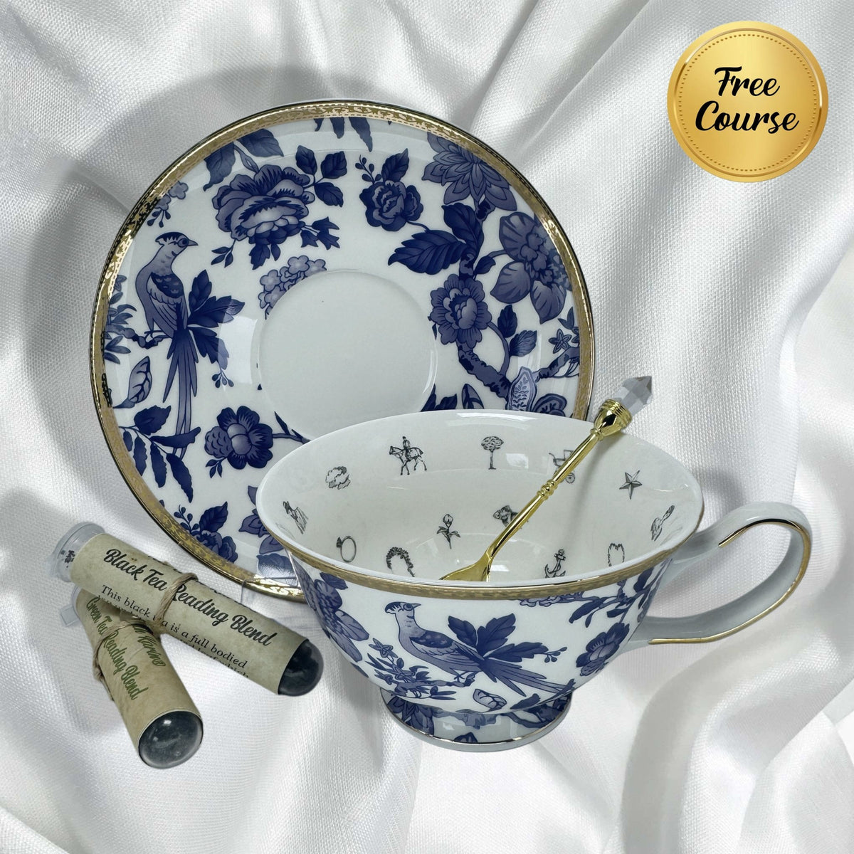 Blue and white teacup set with gold trim, gold teaspoon, and two black tea sample tubes on white satin.