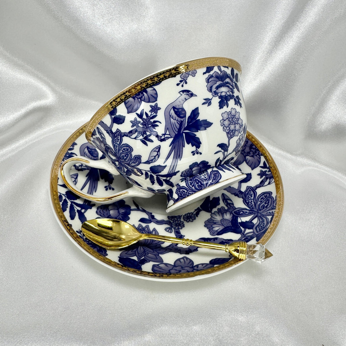 Teacup and saucer set with detailed blue and white bird and flower pattern and gold rim, displayed on a white silk background.