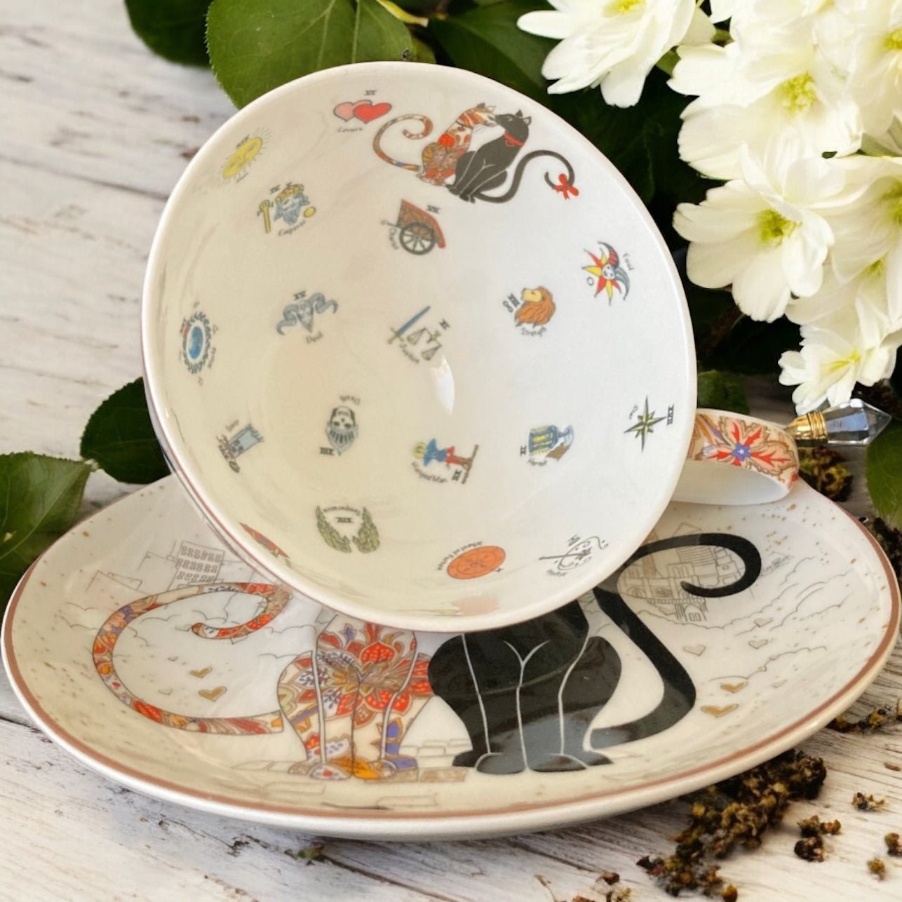 Teacup and saucer with cat illustrations on a wooden surface with flowers in the background