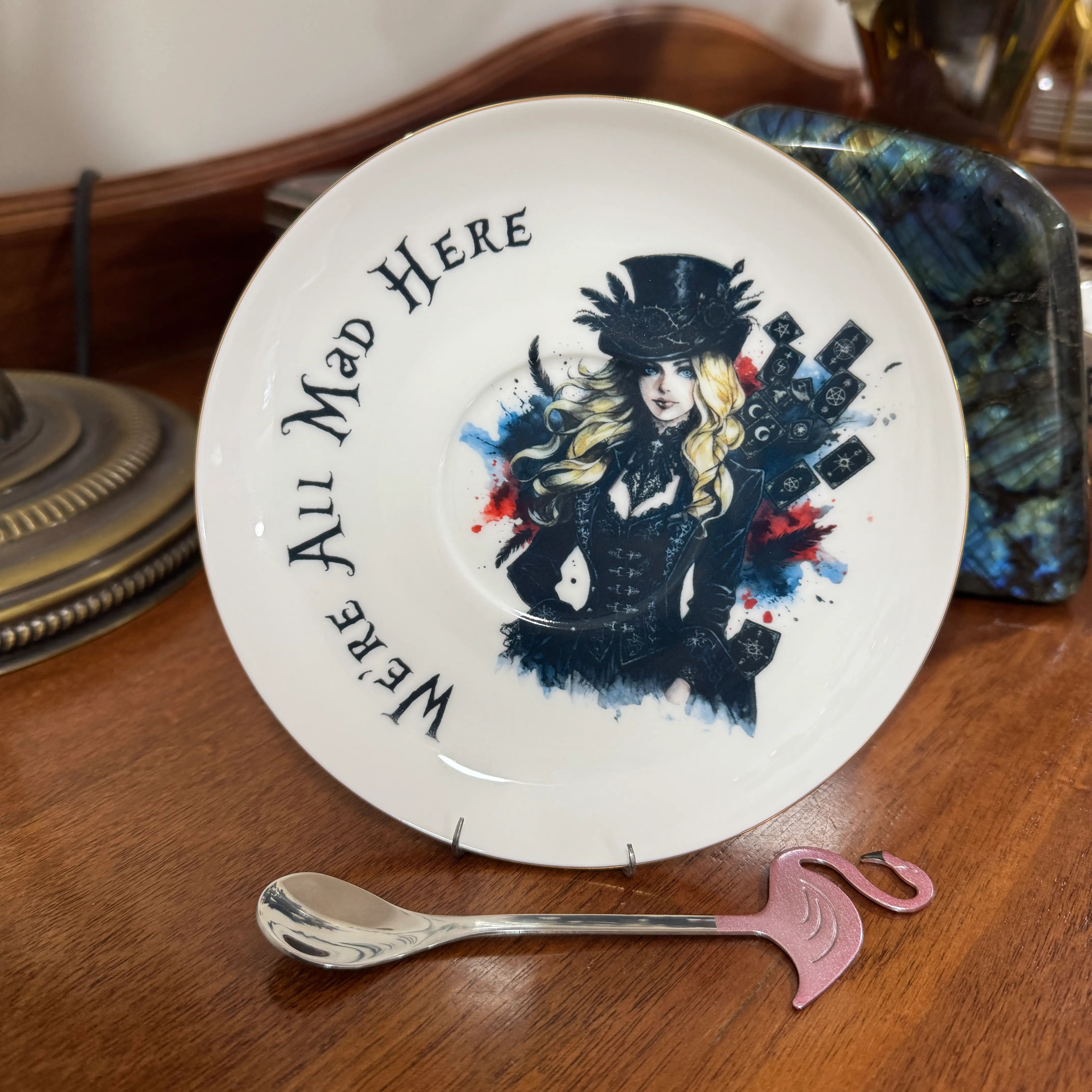 Gothic Alice saucer with feathered top hat, tarot cards and We're All Mad Here lettering, with pink flamingo teaspoon