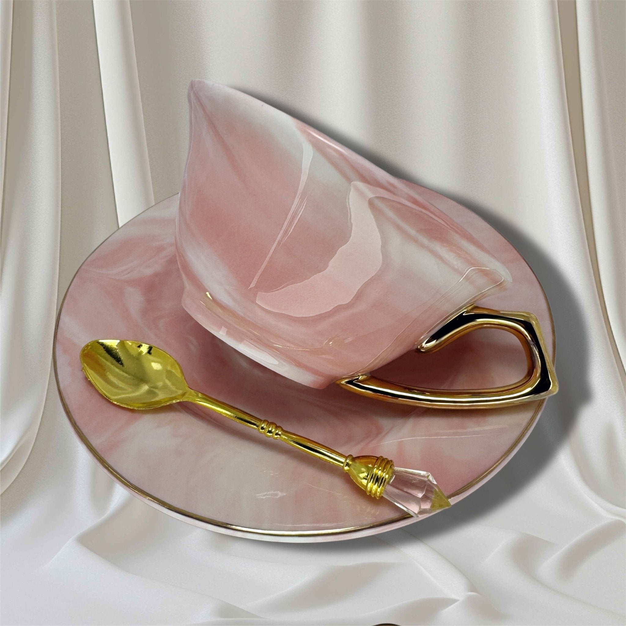 Pink marbled teacup with a high-shine gold handle and gold rim sitting on the matching saucer with a gold spoon resting inside the cup.