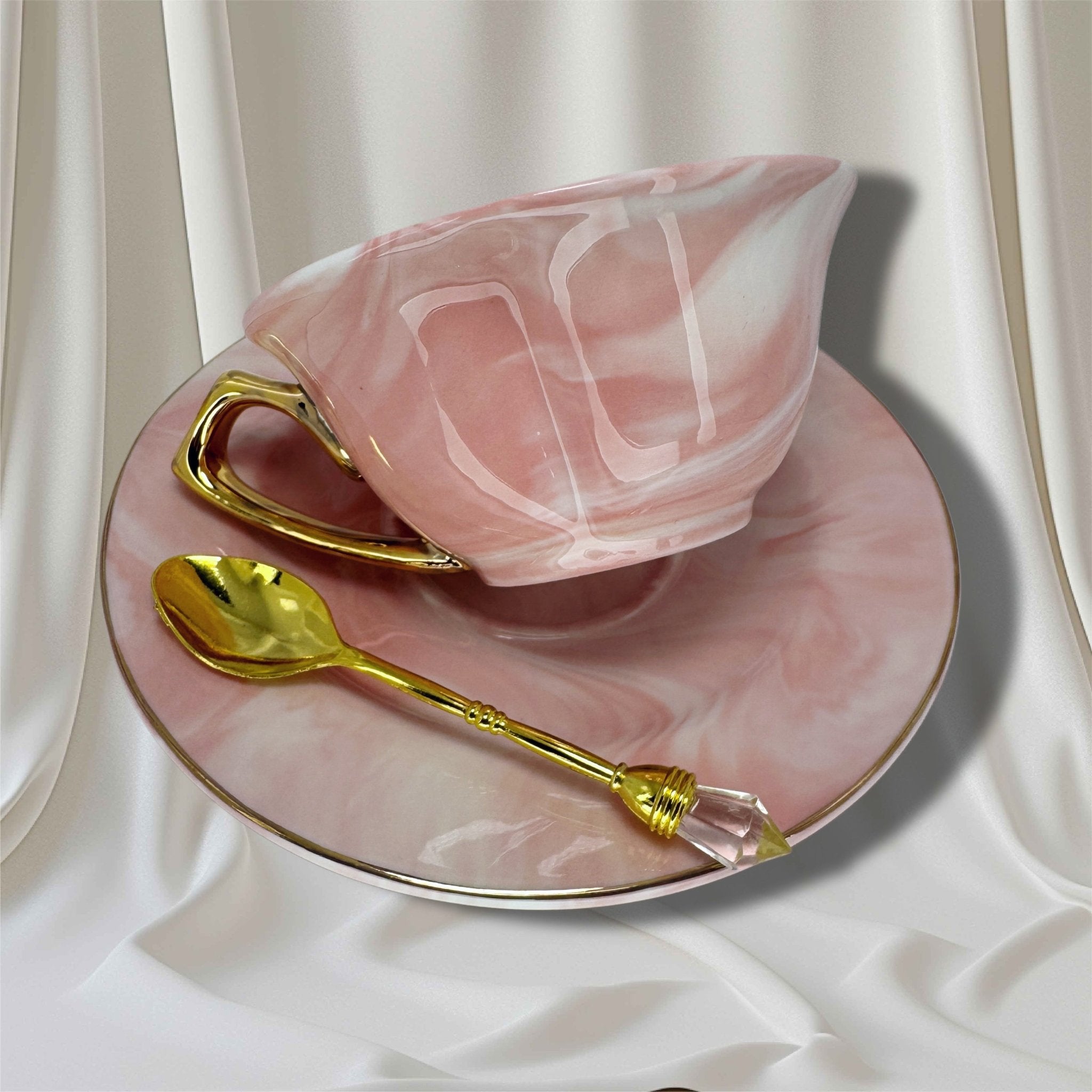 Pink marbled teacup and saucer set with a gold handle, shot from the left side showing the gold spoon resting on the saucer.