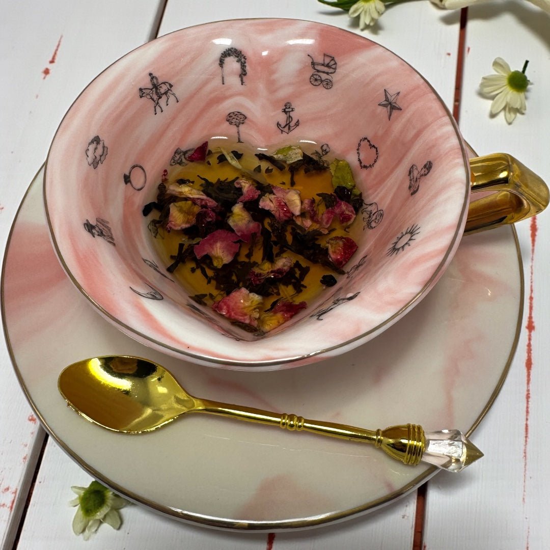 A beautiful pink marble-effect teacup filled with brewing tea, featuring dried rose petals and tea leaves, sitting on a matching saucer with a gold spoon, surrounded by white flowers.
