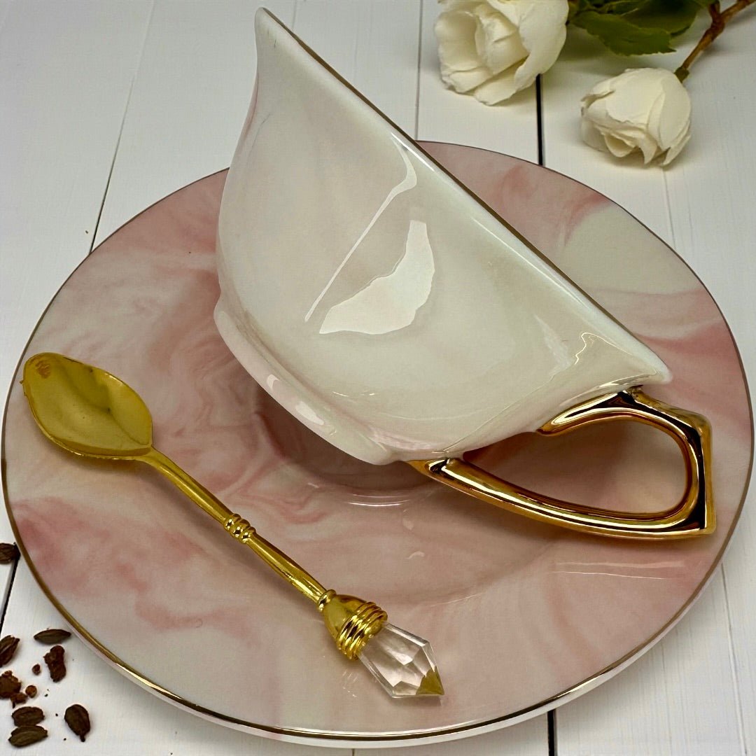 Pink and white marble teacup with gold handle and rim, sitting on its matching saucer, beside a gold spoon and white flowers.