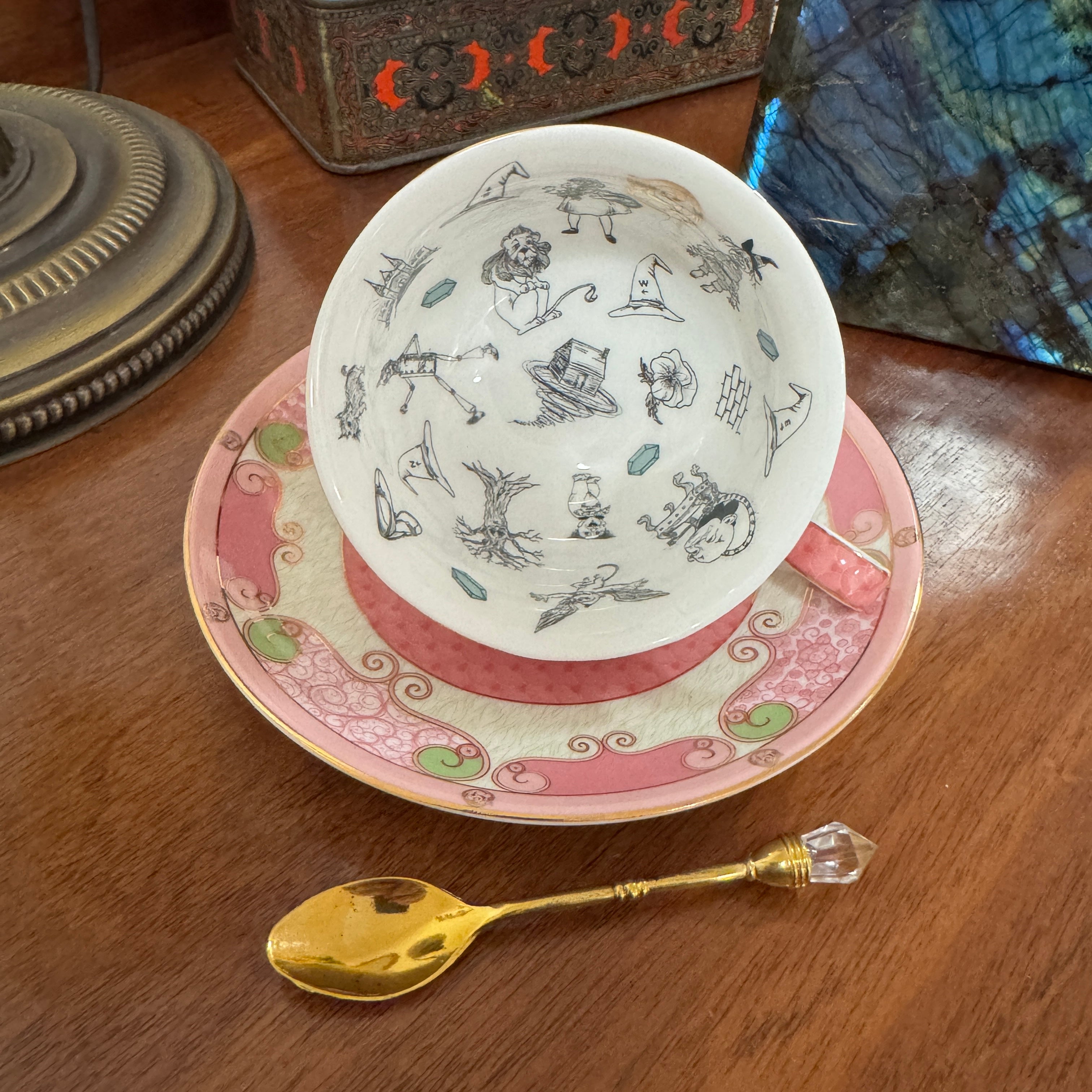 Teacup with decorative design on a saucer, accompanied by a gold spoon on a wooden surface.