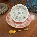 Teacup with decorative design on a saucer, accompanied by a gold spoon on a wooden surface.