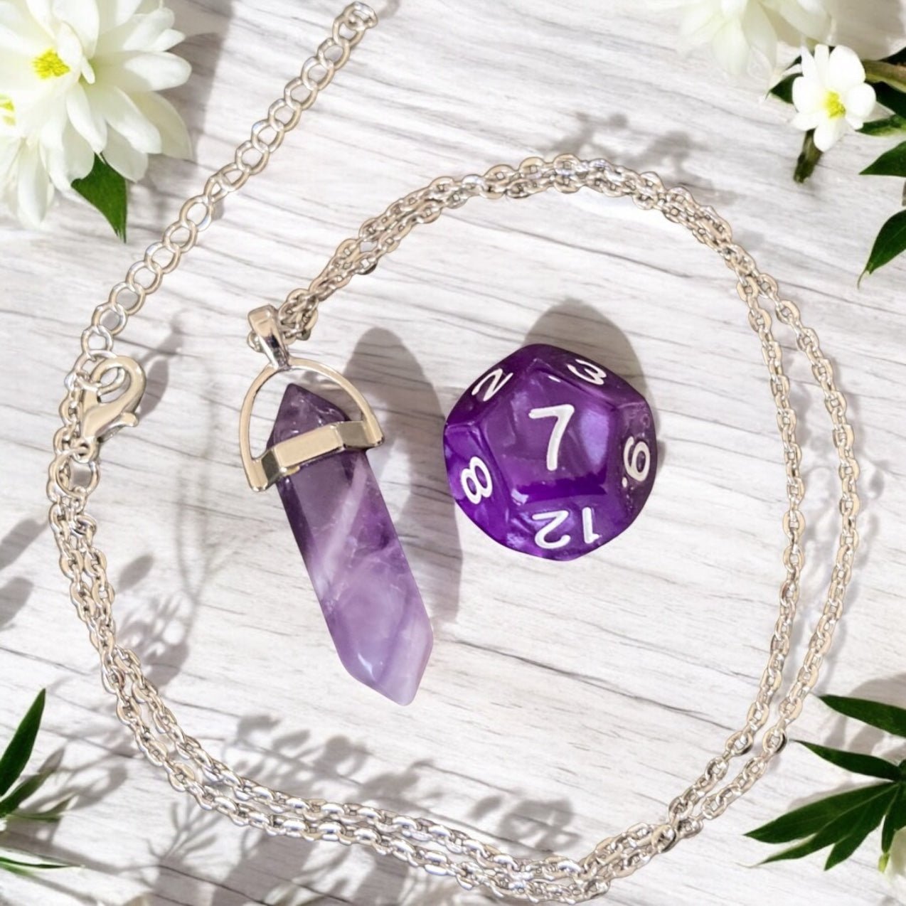 Amethyst crystal necklace with silver chain on a white textured surface with a purple die and flowers.