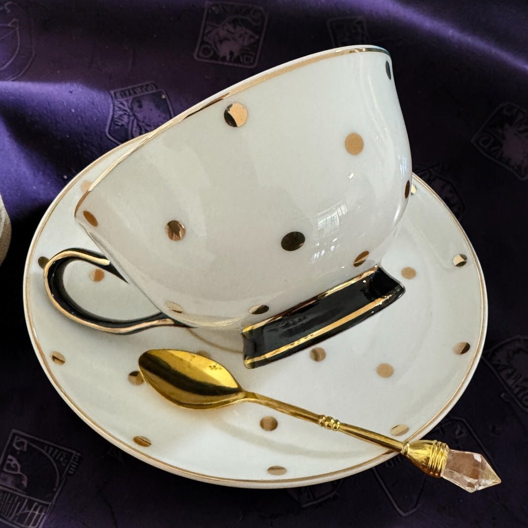 White teacup with gold polka dots on a matching saucer, accompanied by a gold spoon and black handle on a purple fabric background.