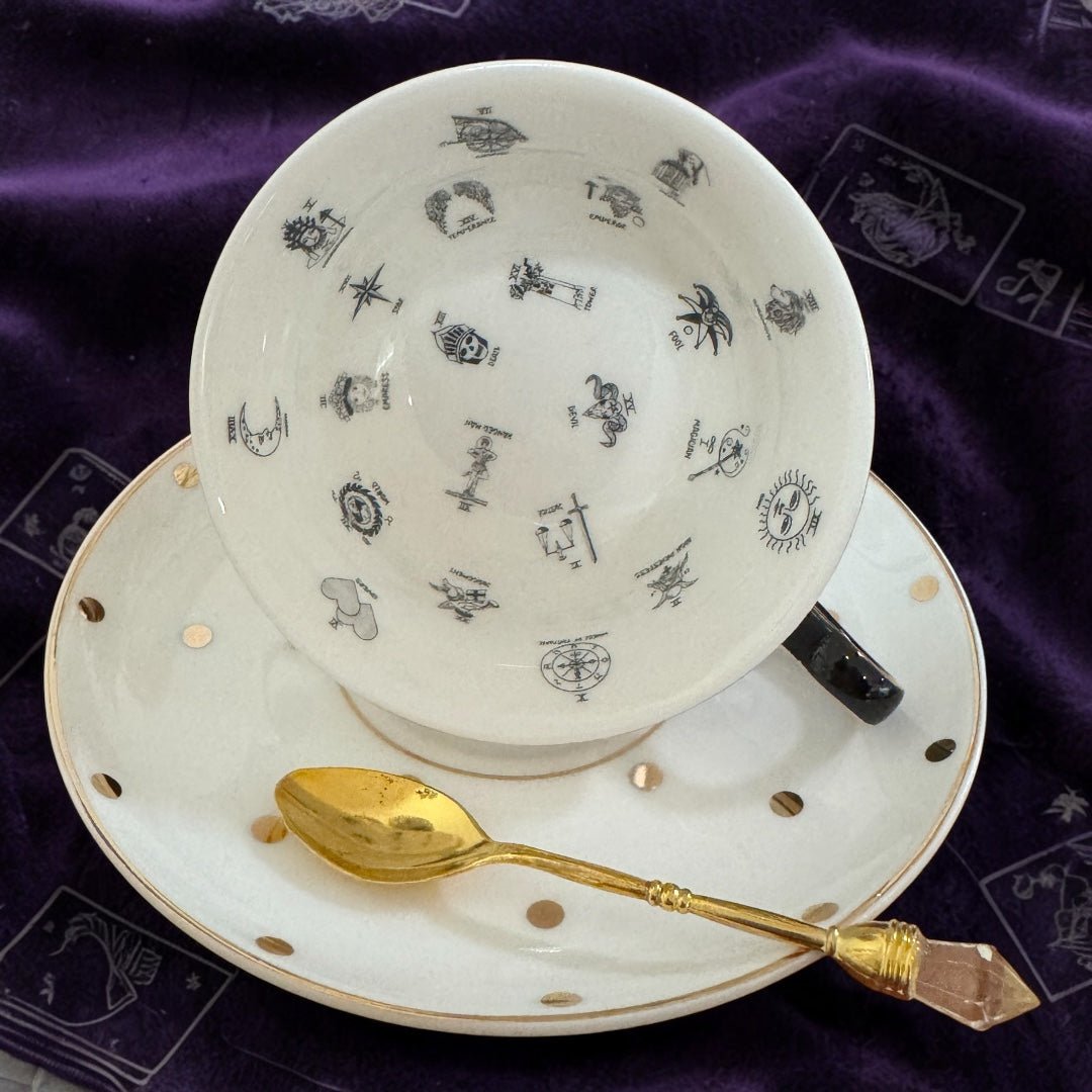 Teacup with decorative patterns on a matching saucer and a gold spoon on a purple fabric background.